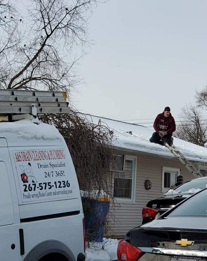 Person on snowy roof removing ice with ladder. Service van parked in front of a house.