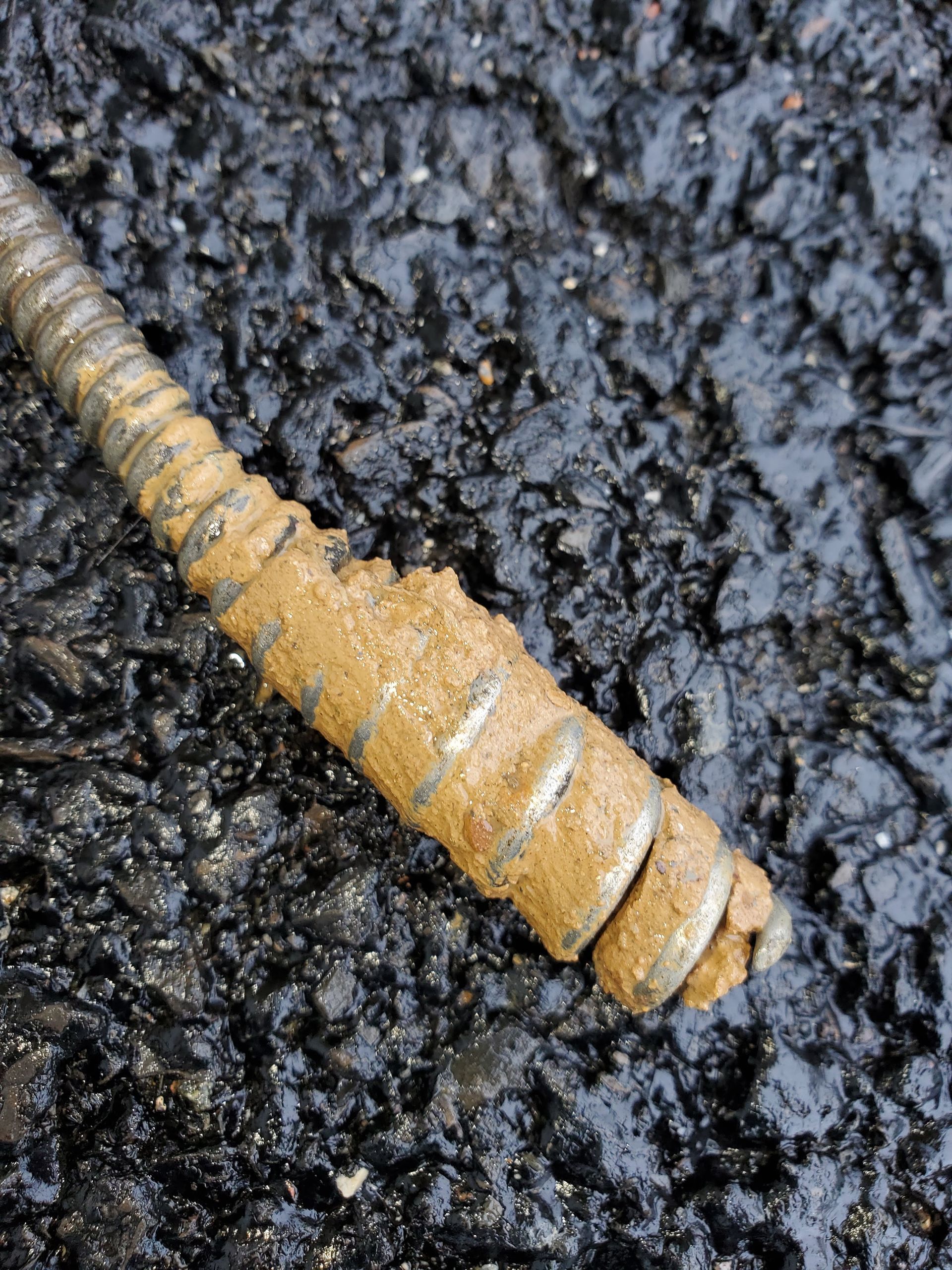 A muddy metal rod lies on asphalt. The rod's spiral end is covered in light brown mud.