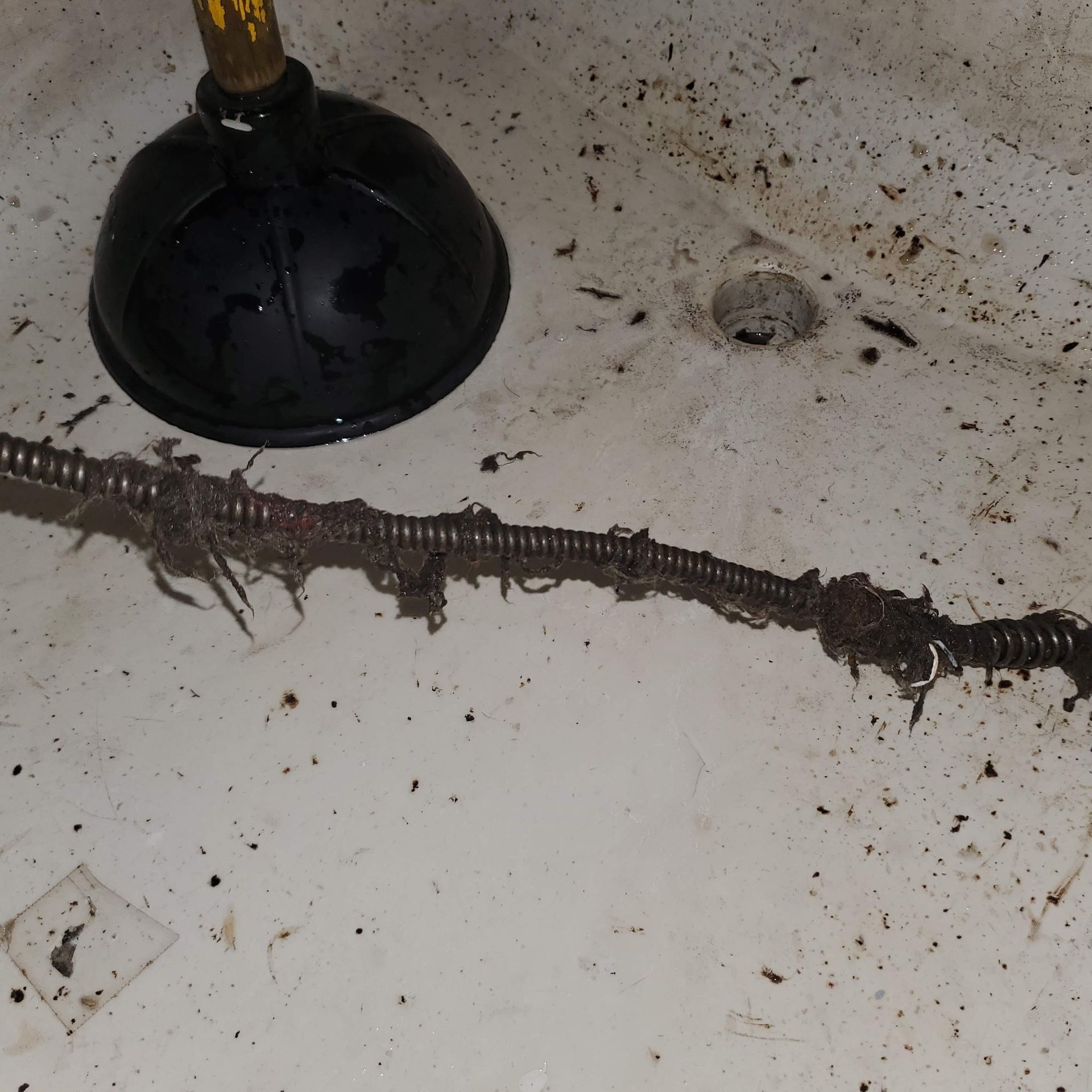A drain snake and plunger next to a bathtub drain with debris.