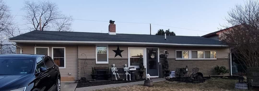A one-story beige house with dark trim. A black star hangs on the wall, and a black car is parked in the driveway.