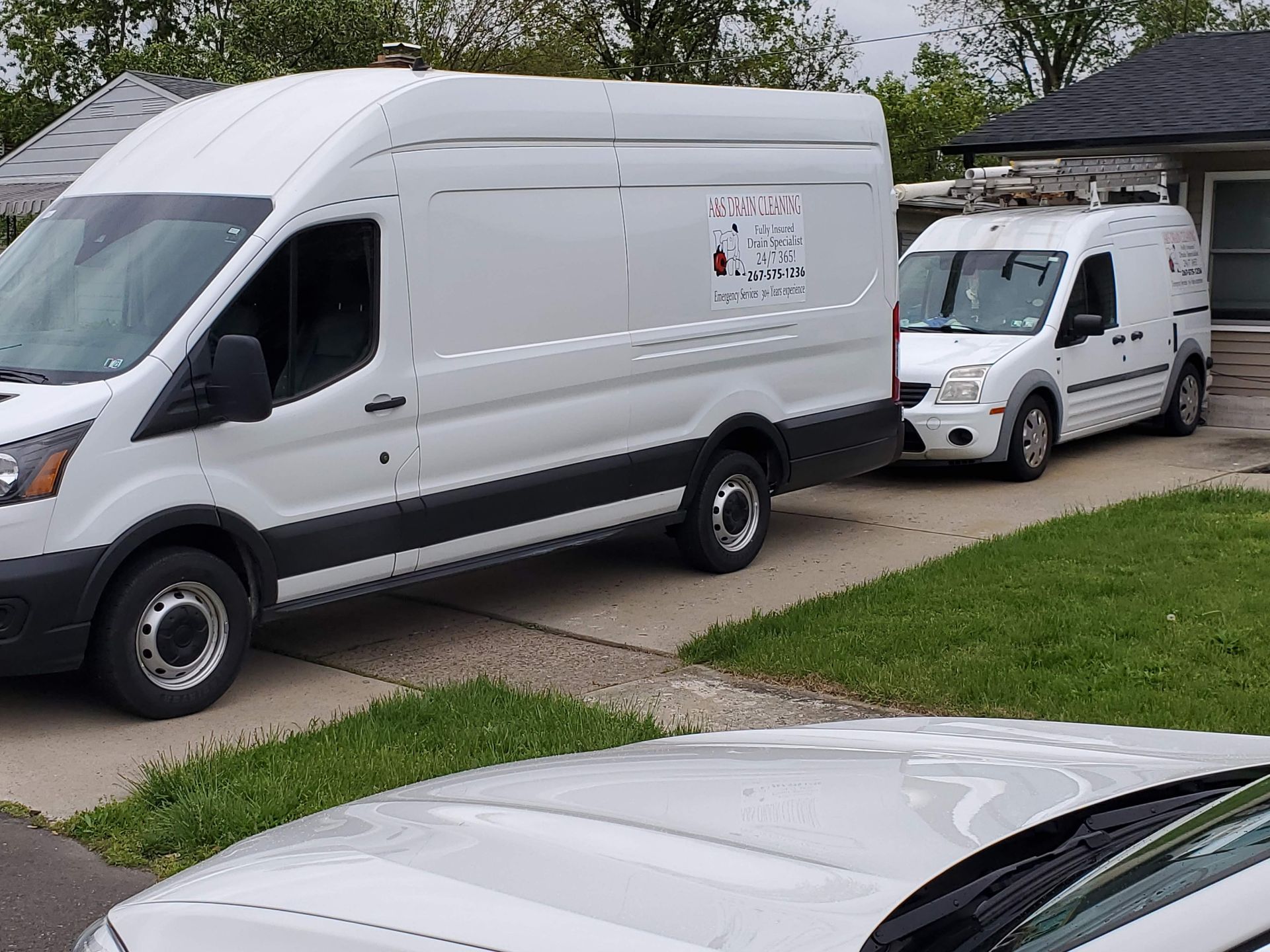 Two white work vans parked in a driveway next to a house; one is much larger than the other.