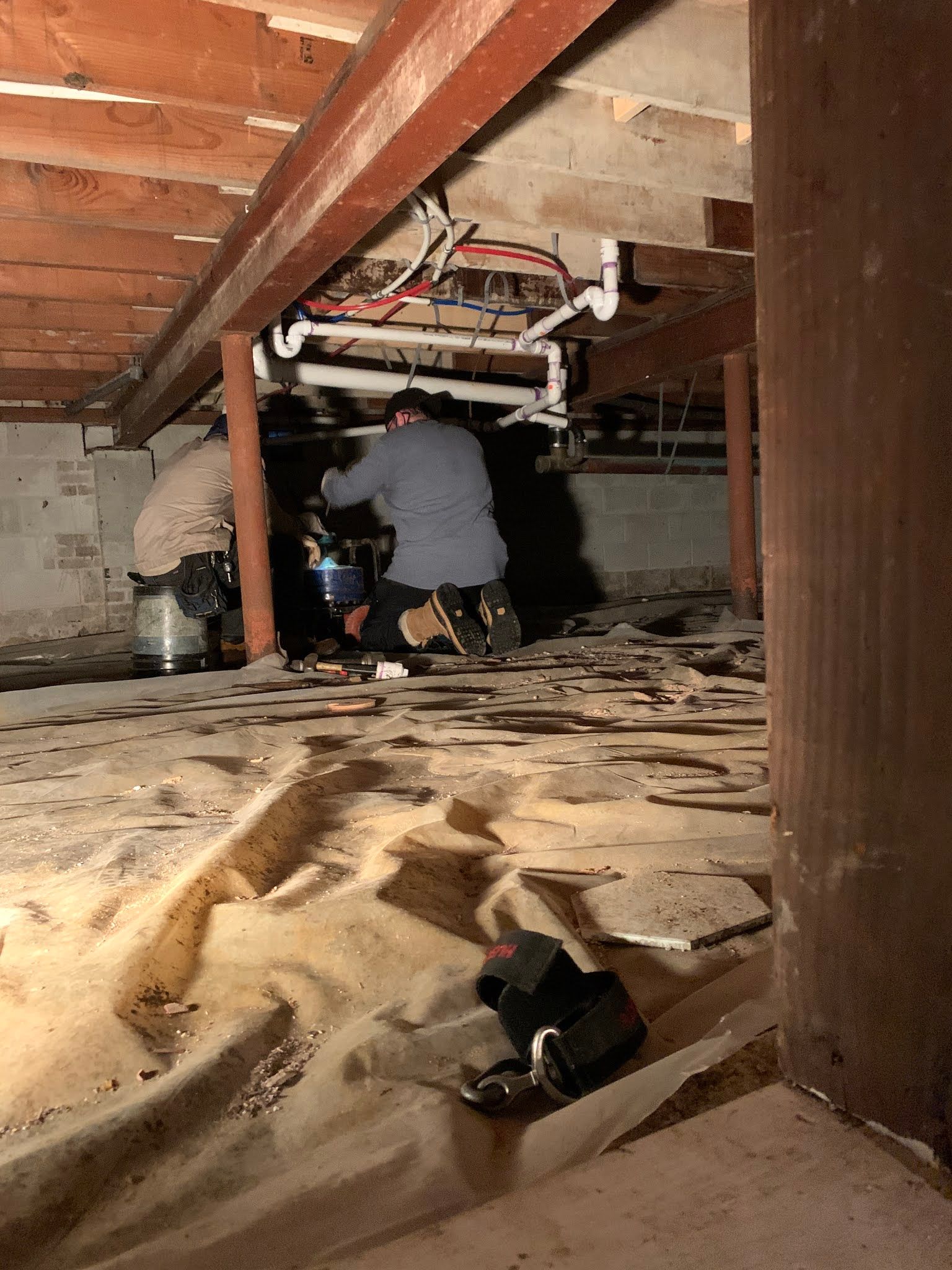 Two people working under a building with plumbing and tools. Brown beams, pipes, and white plastic covering.