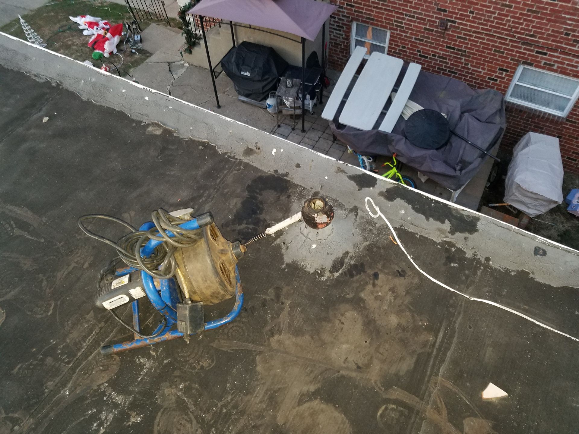 Overhead view of a black roof with equipment and a sitting man; adjacent to a patio with furniture and a brick wall.