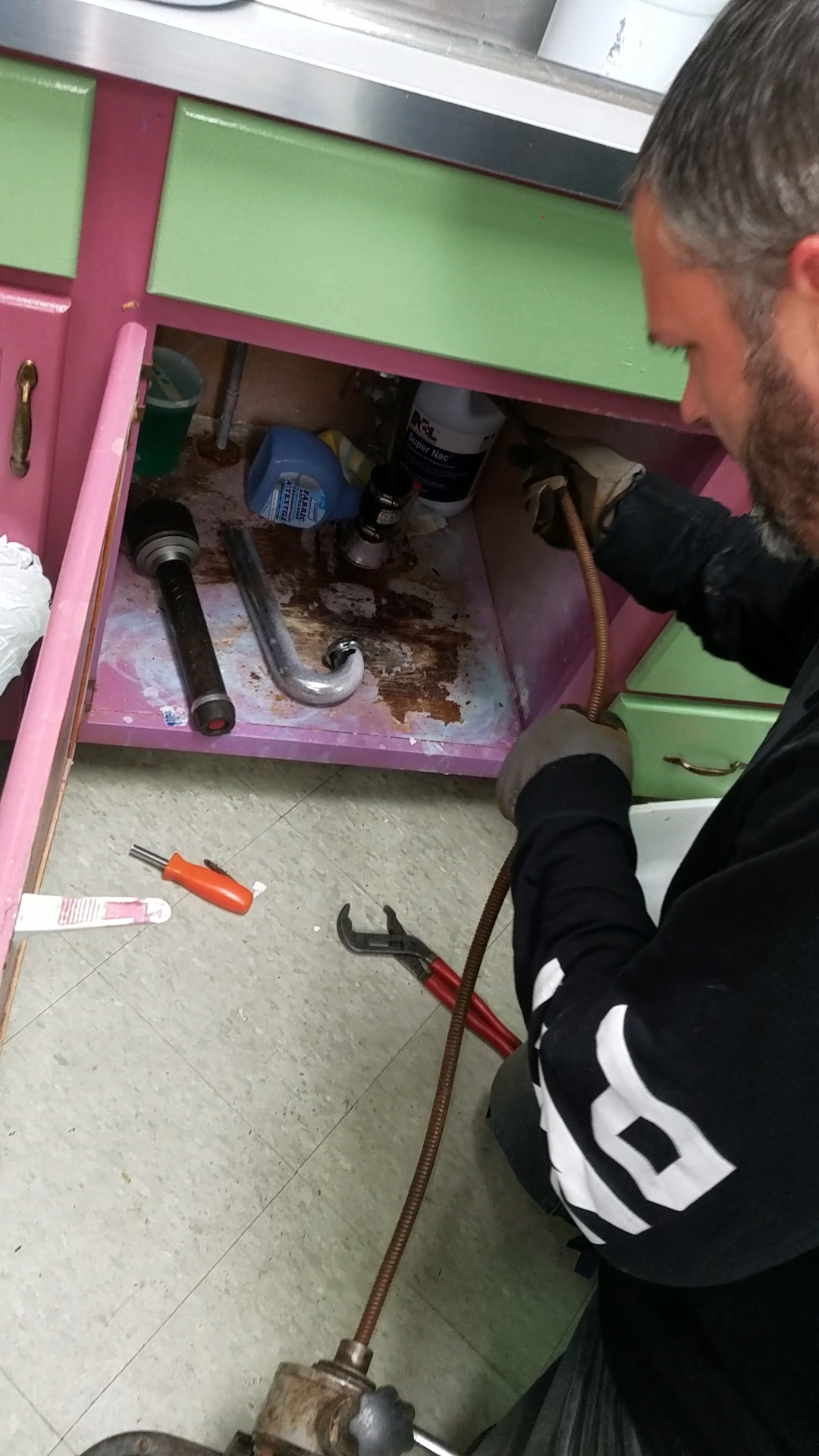 Man working under a pink and green kitchen sink, using a drain snake.