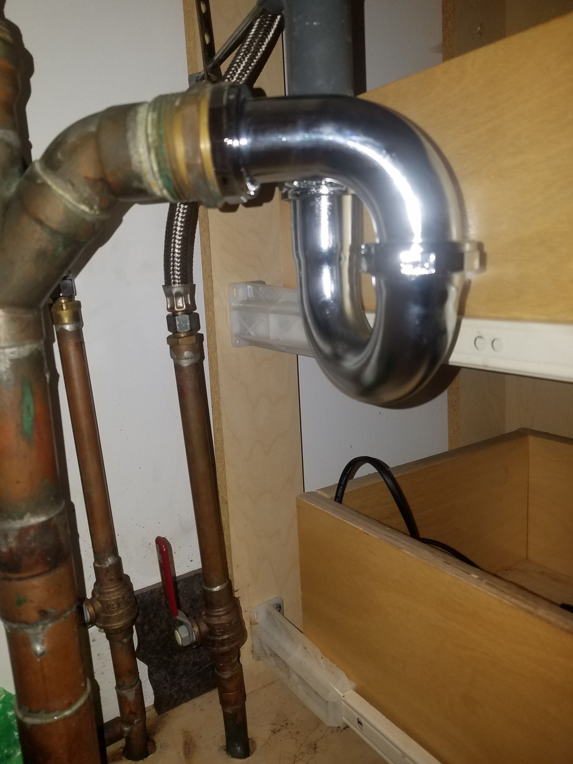 Plumbing under a sink: shiny chrome P-trap connected to copper pipes and wood cabinetry.