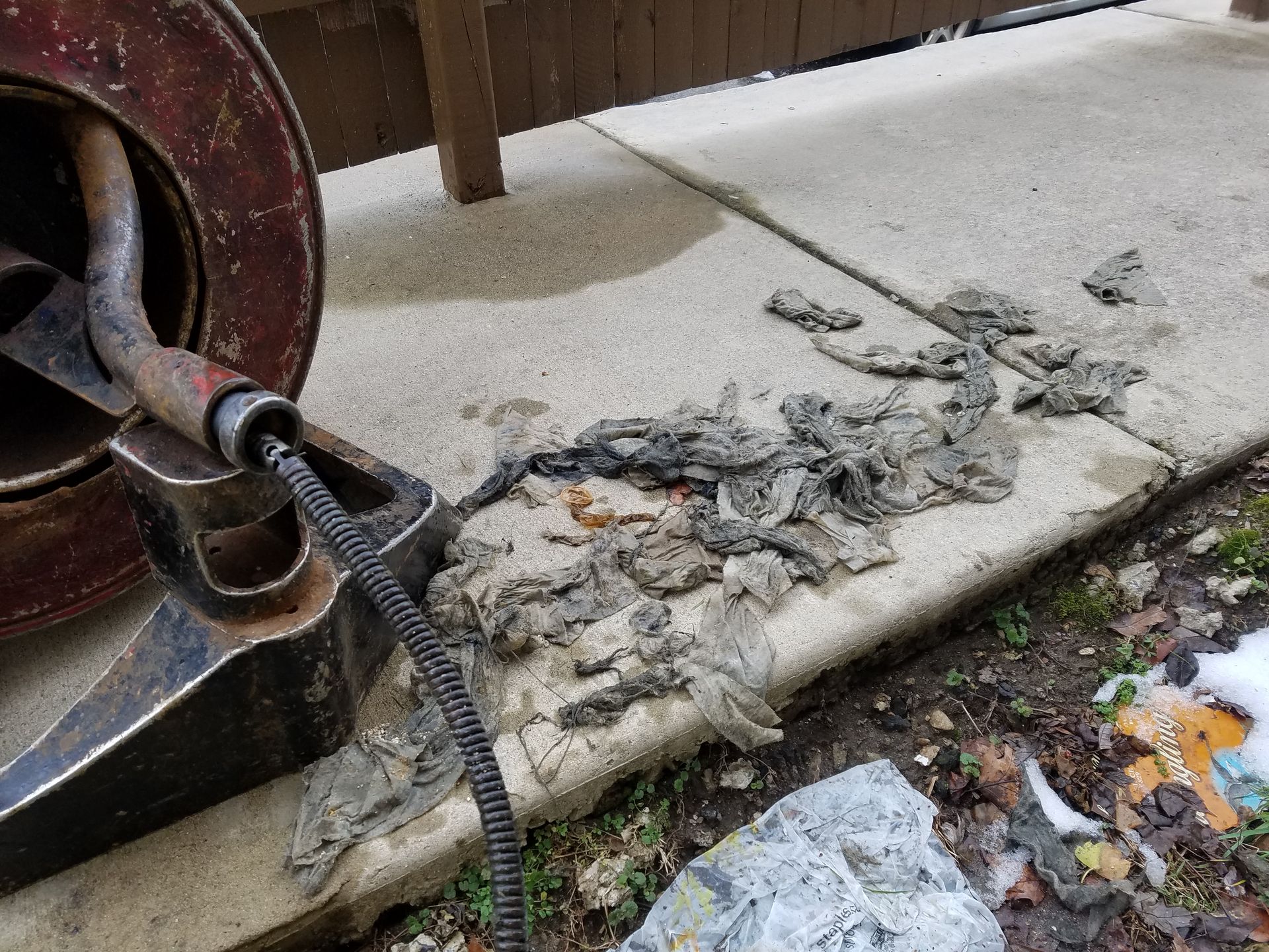 A drain snake unearths a pile of debris on a concrete sidewalk near a building.