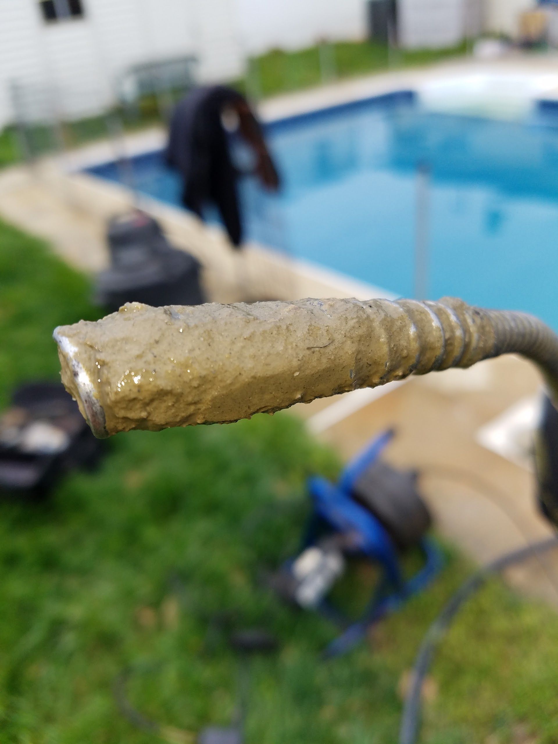Close-up of a dirty, metal drain snake tip, with a pool and person in the background.