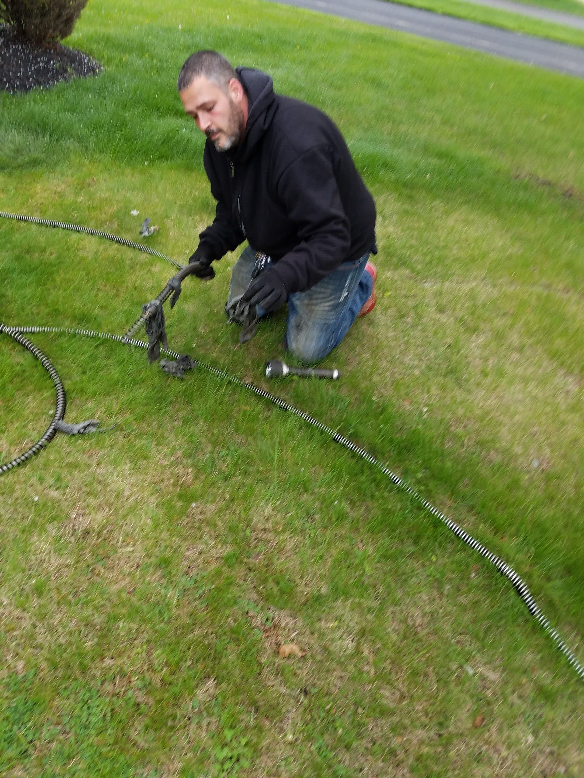 Man kneeling on grass, working on metal chains. He wears a black jacket, gloves, and jeans.