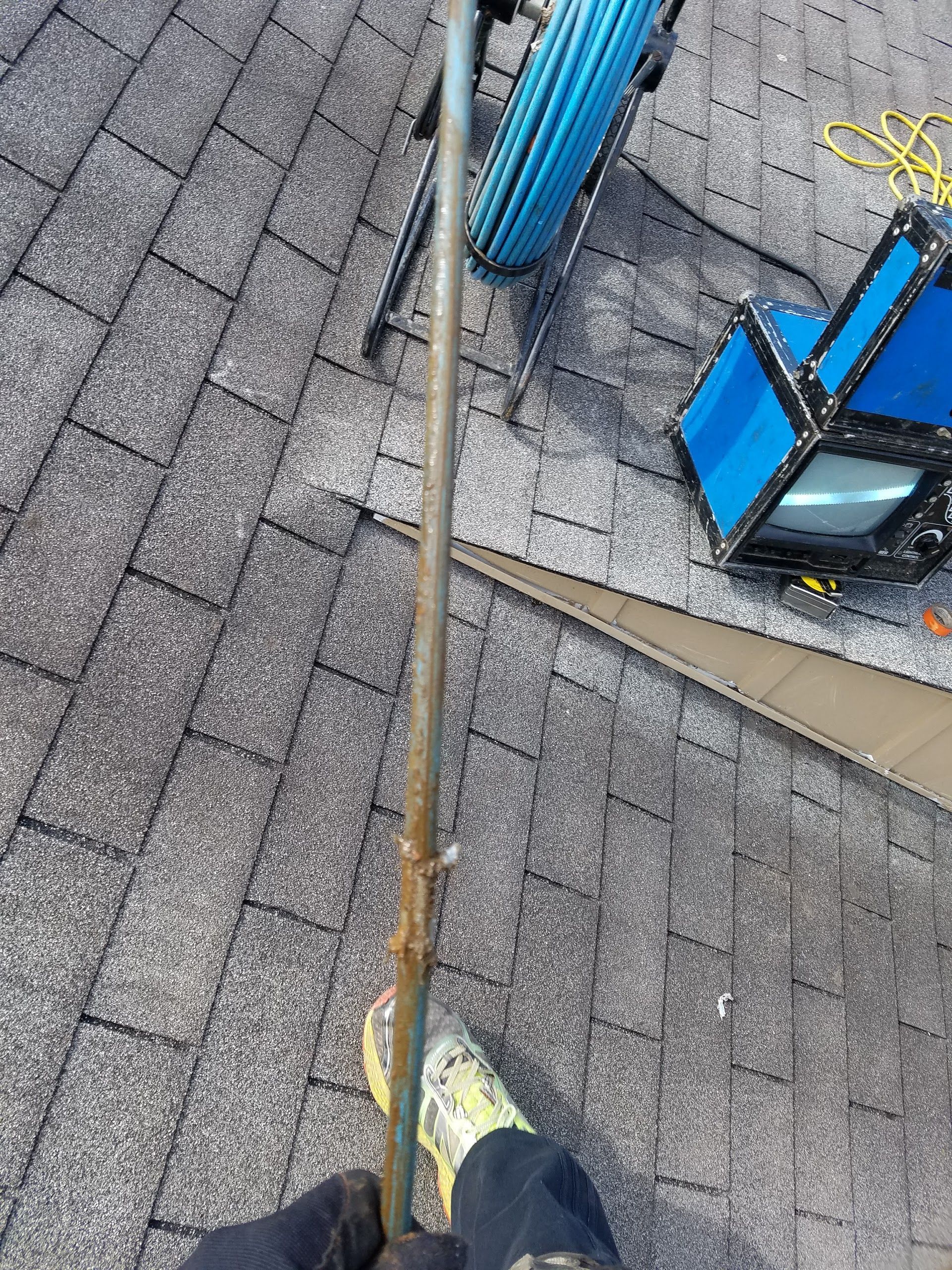 Person on a roof holding a pole, looking down.  Shingles, equipment, and cable in the background.
