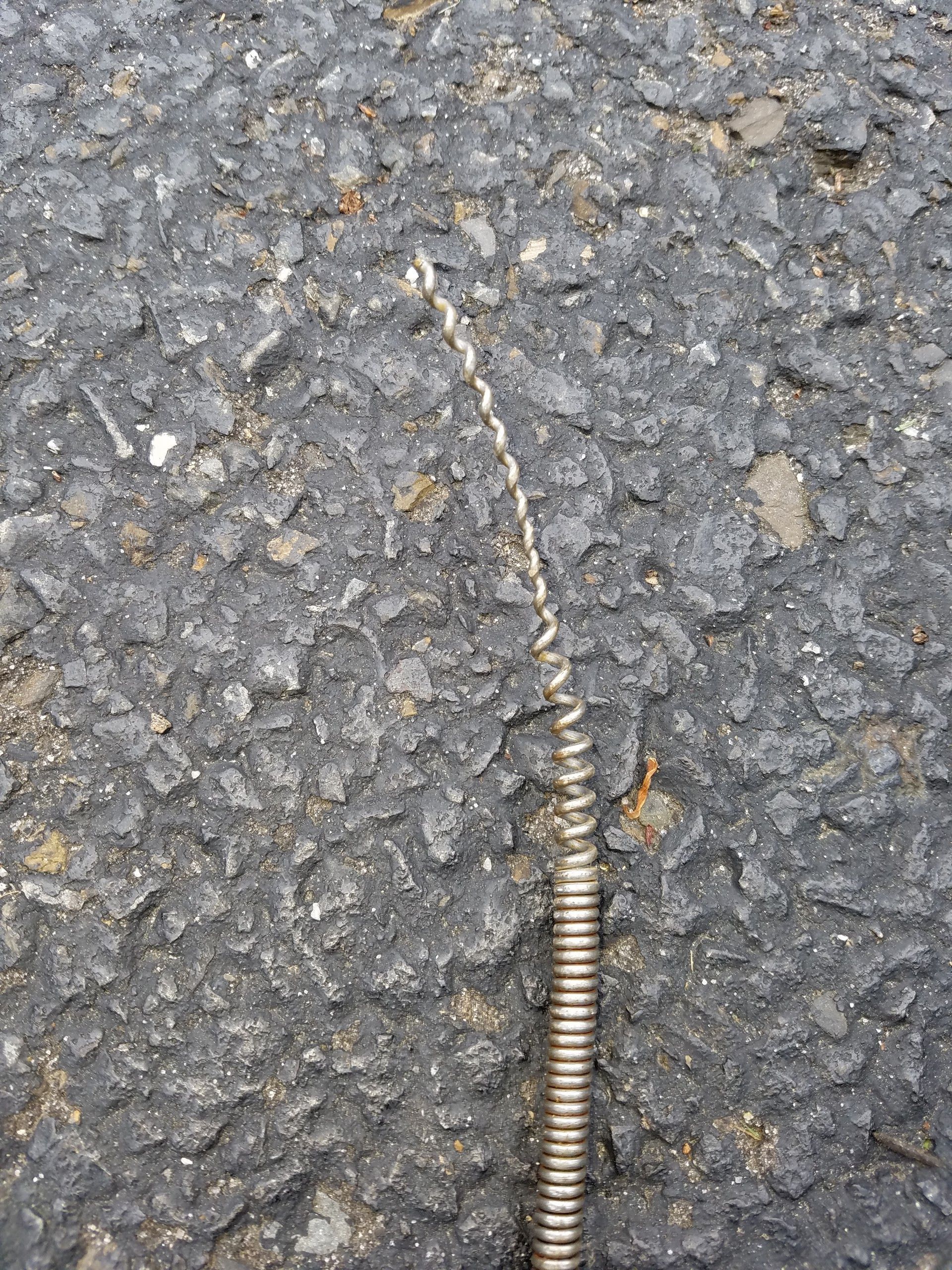 A thin, metal, spiral-shaped drain snake lying on dark asphalt.