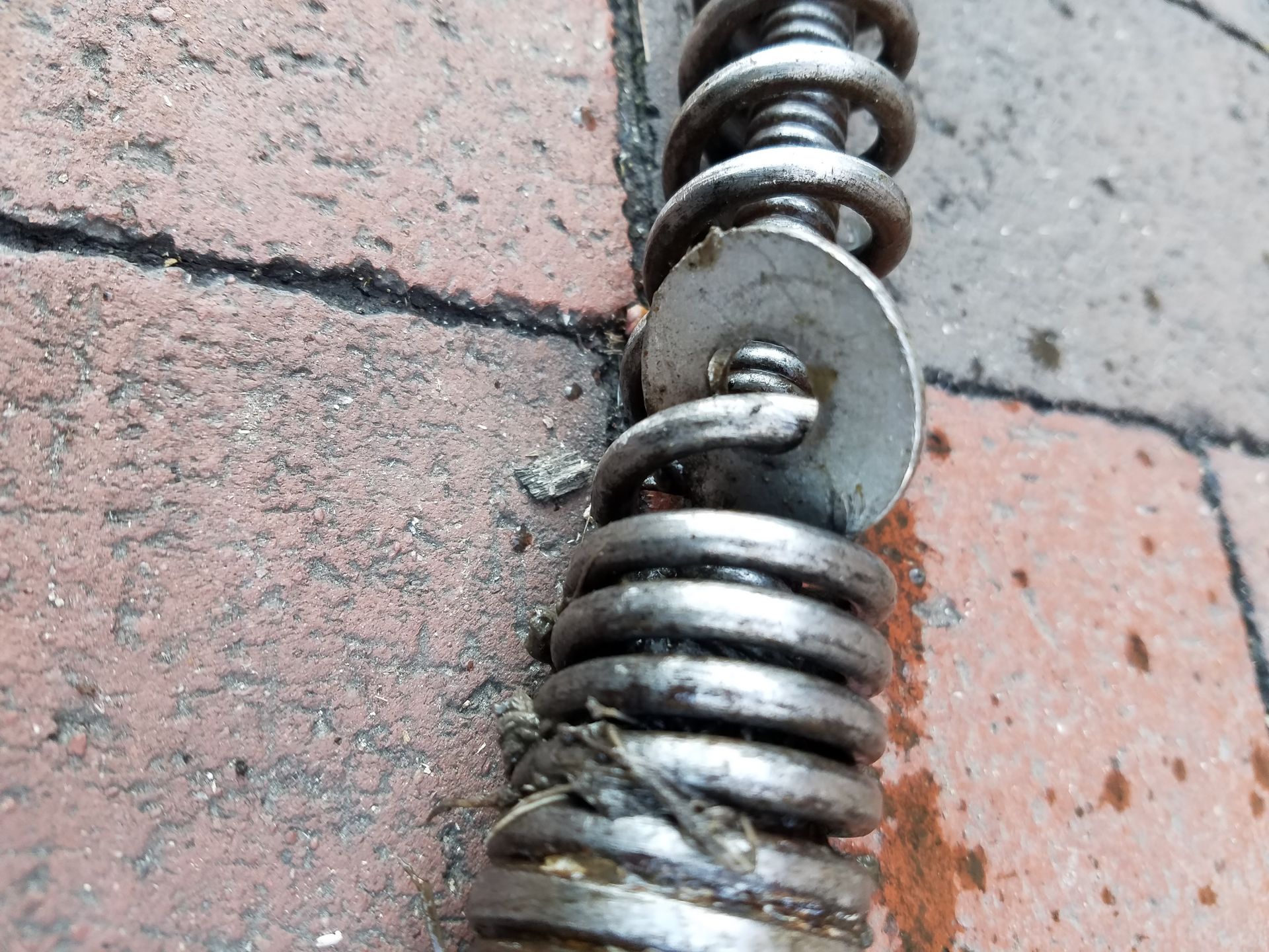 Close-up of a metal drain snake lying on brick. A metal disc is attached to the coiled spring.