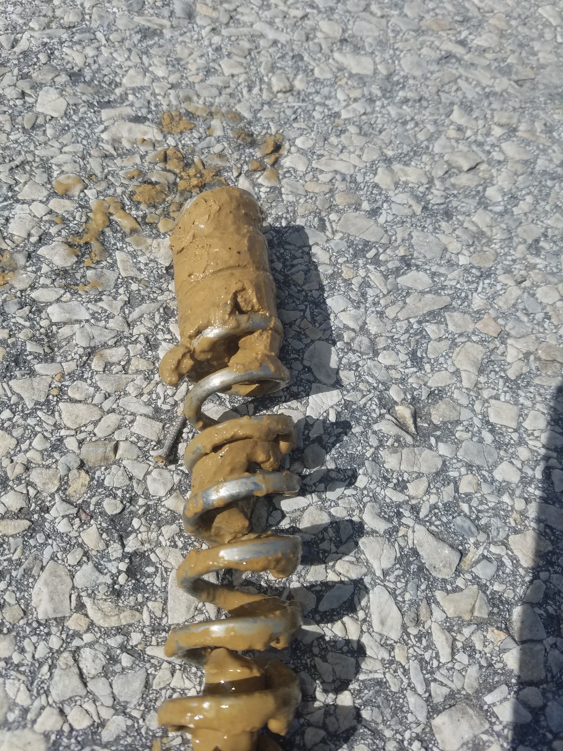 Soil sample clinging to a metal auger, resting on a paved surface.