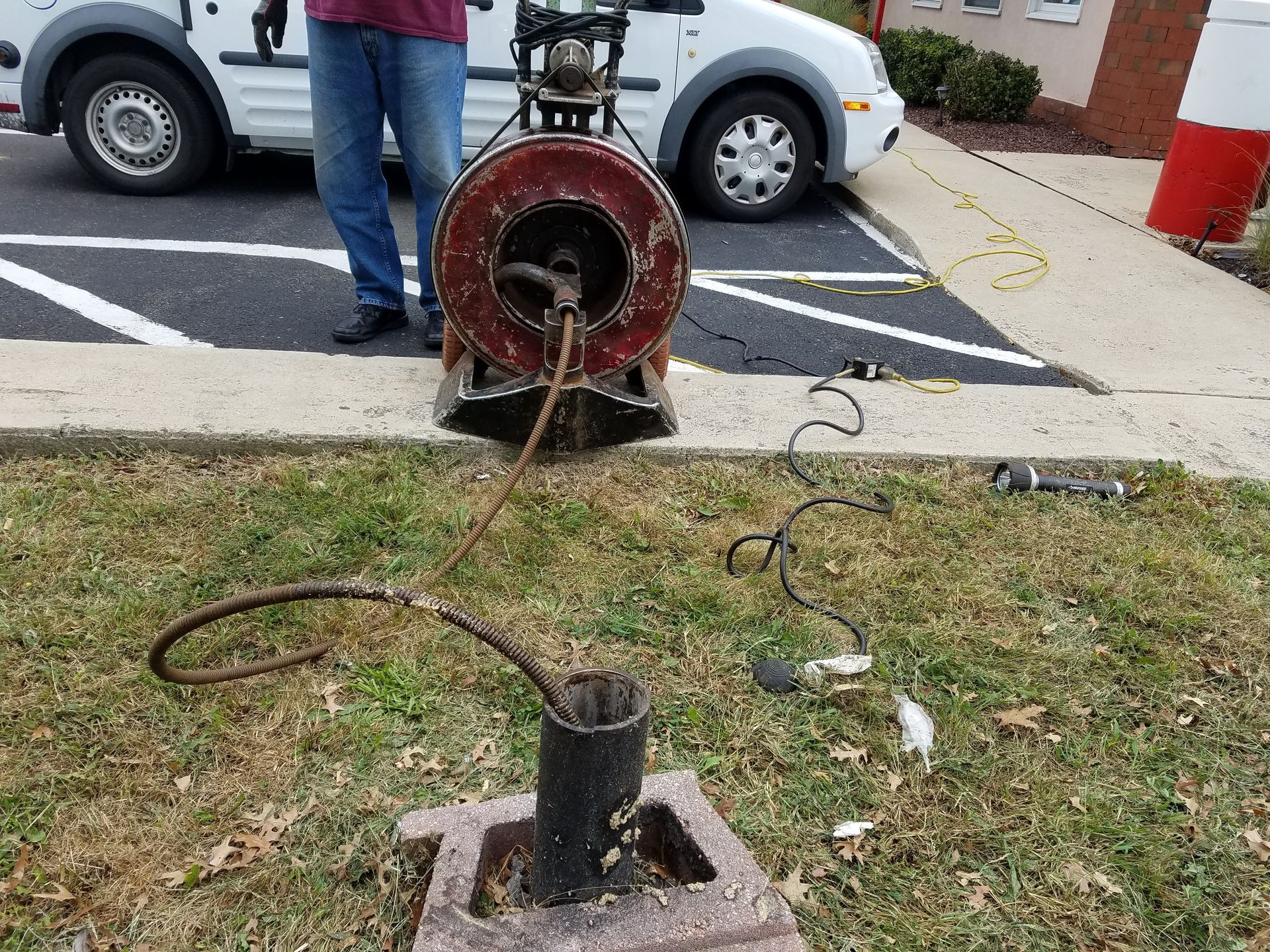 Man using a drain cleaning machine near a concrete block and parked vehicles.