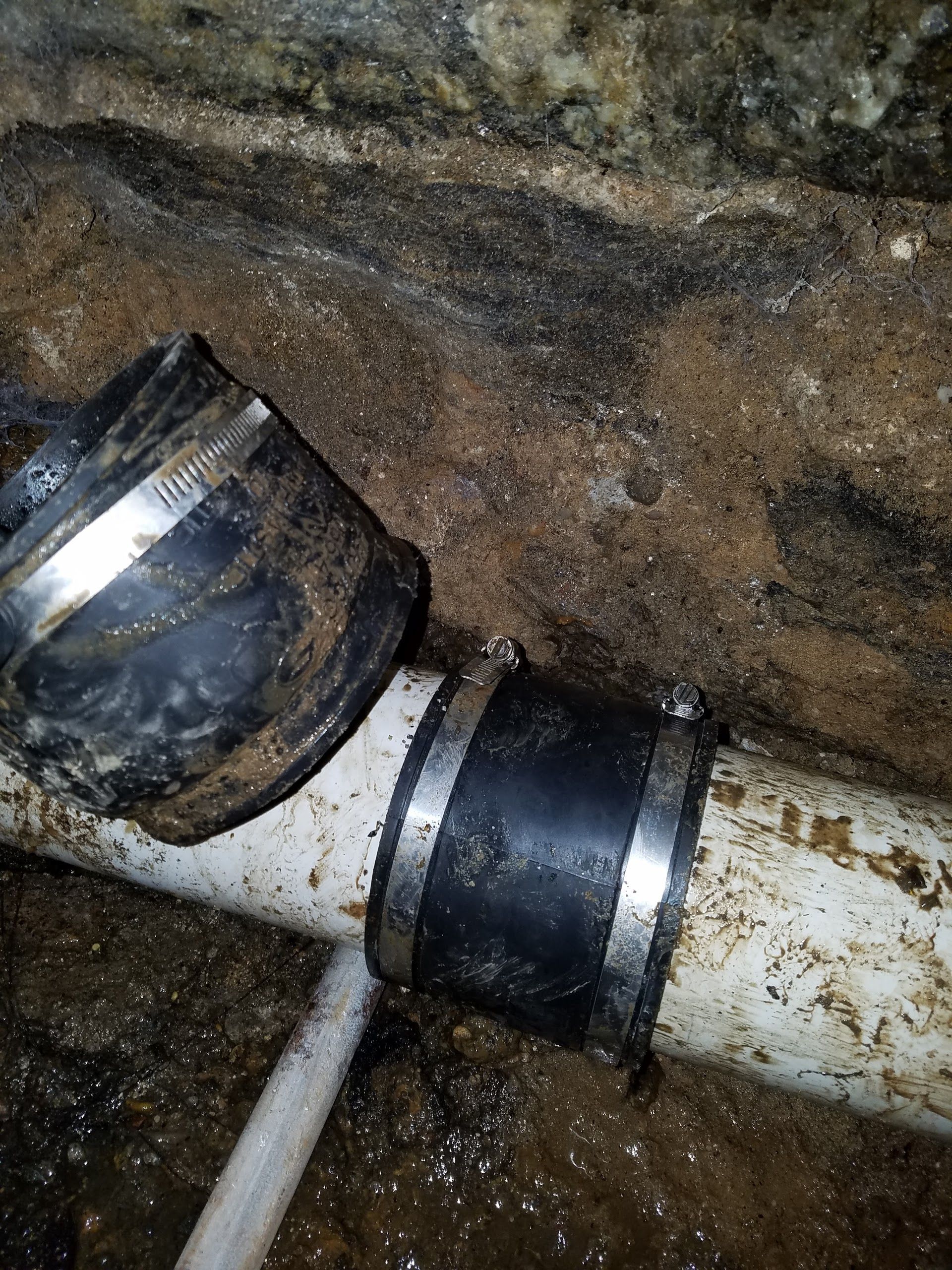 Two black rubber couplings connect white PVC pipes in a dirty, dark setting.