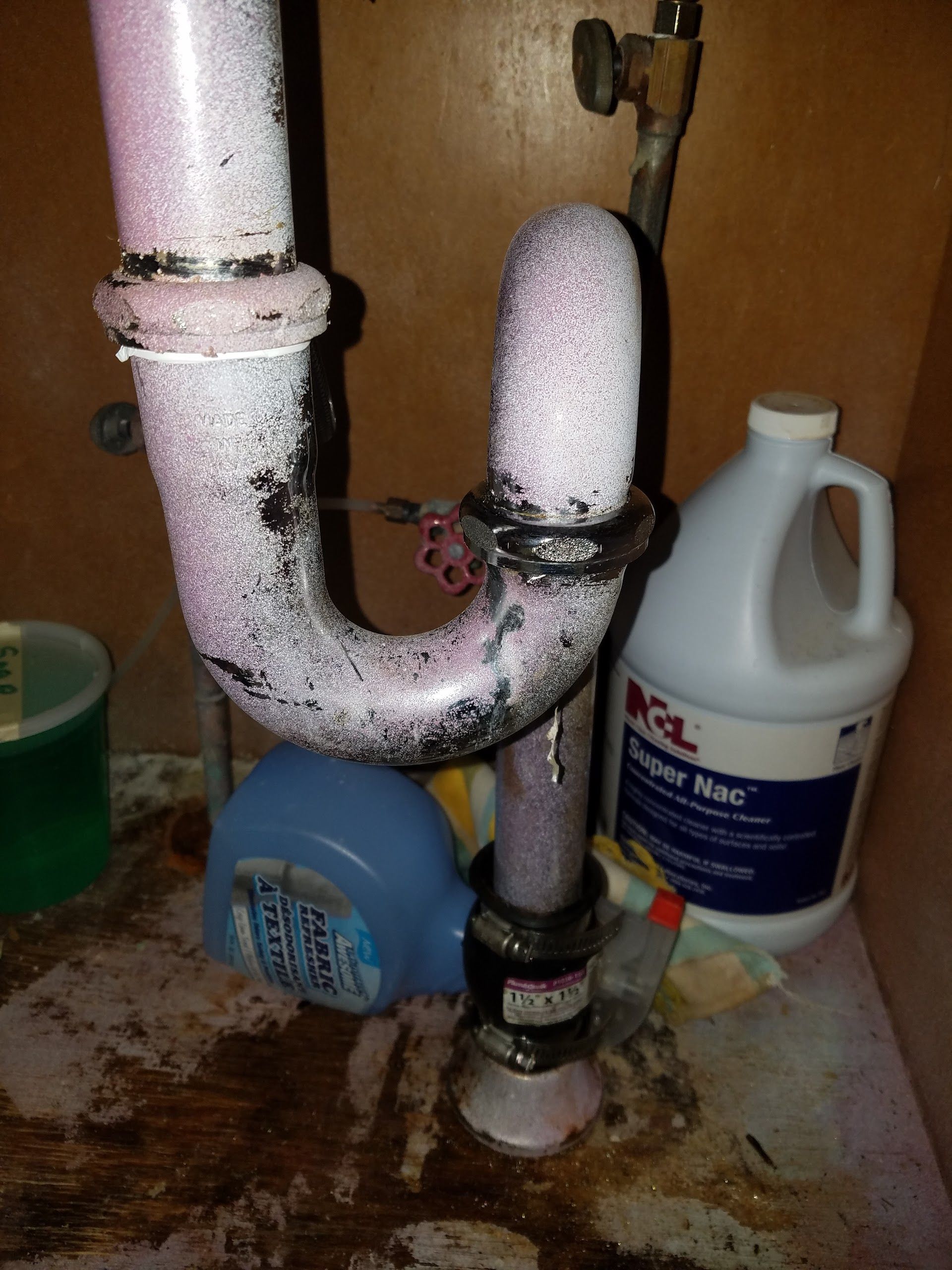 Under-sink plumbing with pink-ish residue and cleaning supplies, beneath a cabinet.