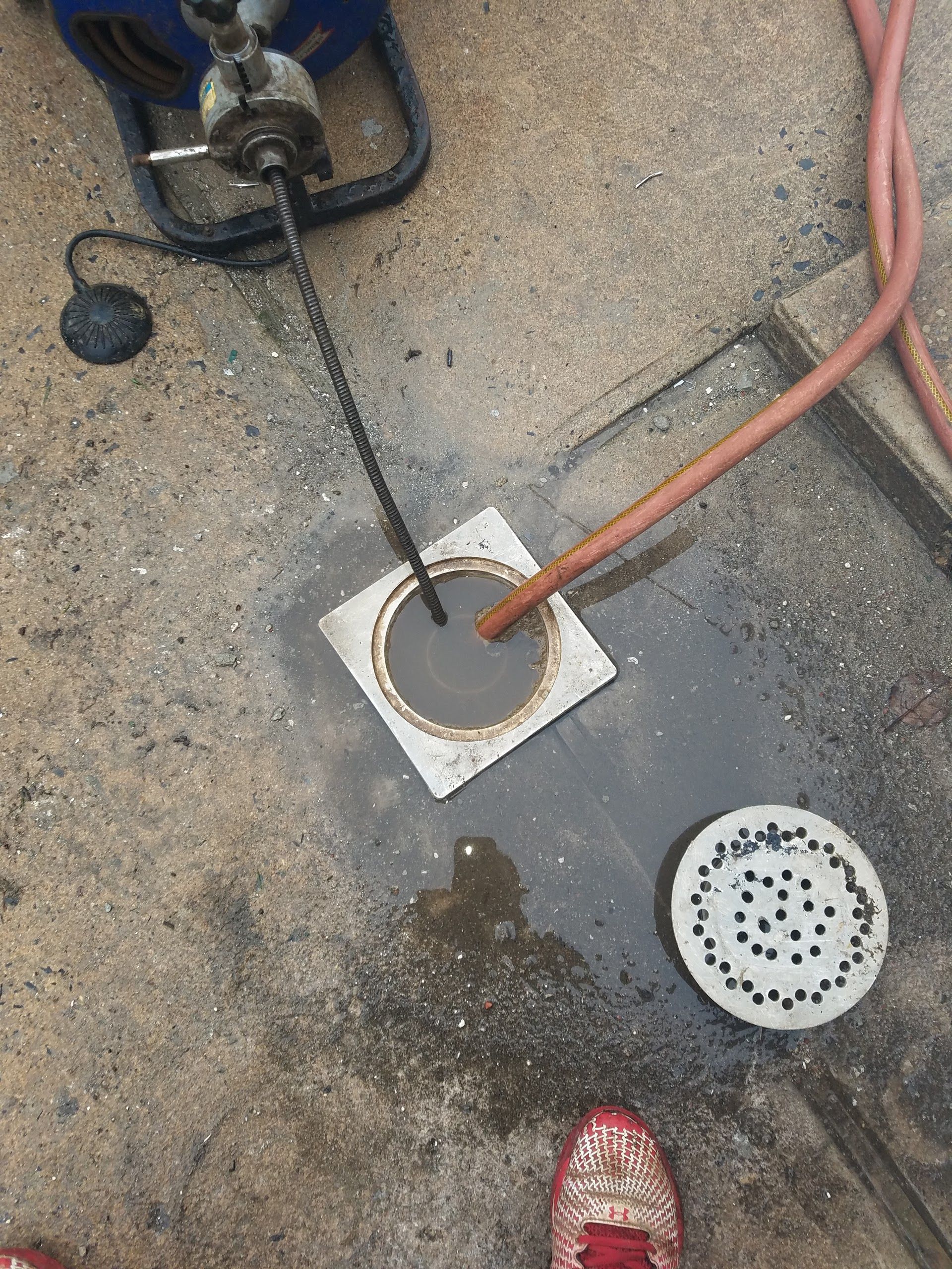 A drain is being unclogged with a snake tool. Water and tools on concrete.