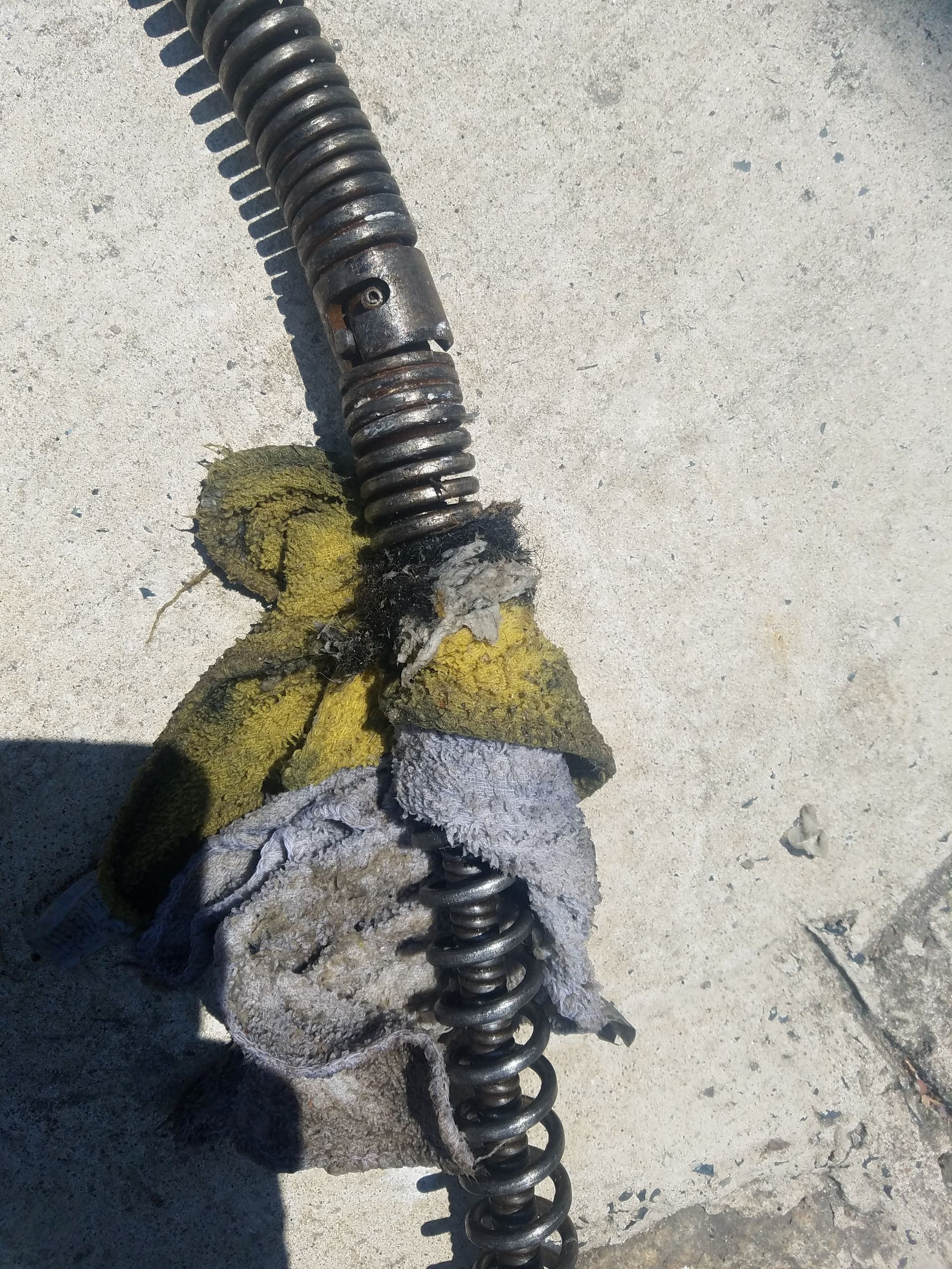A dirty, flexible drain snake with dark oil residue is wrapped with a yellow and gray rag on a concrete surface.
