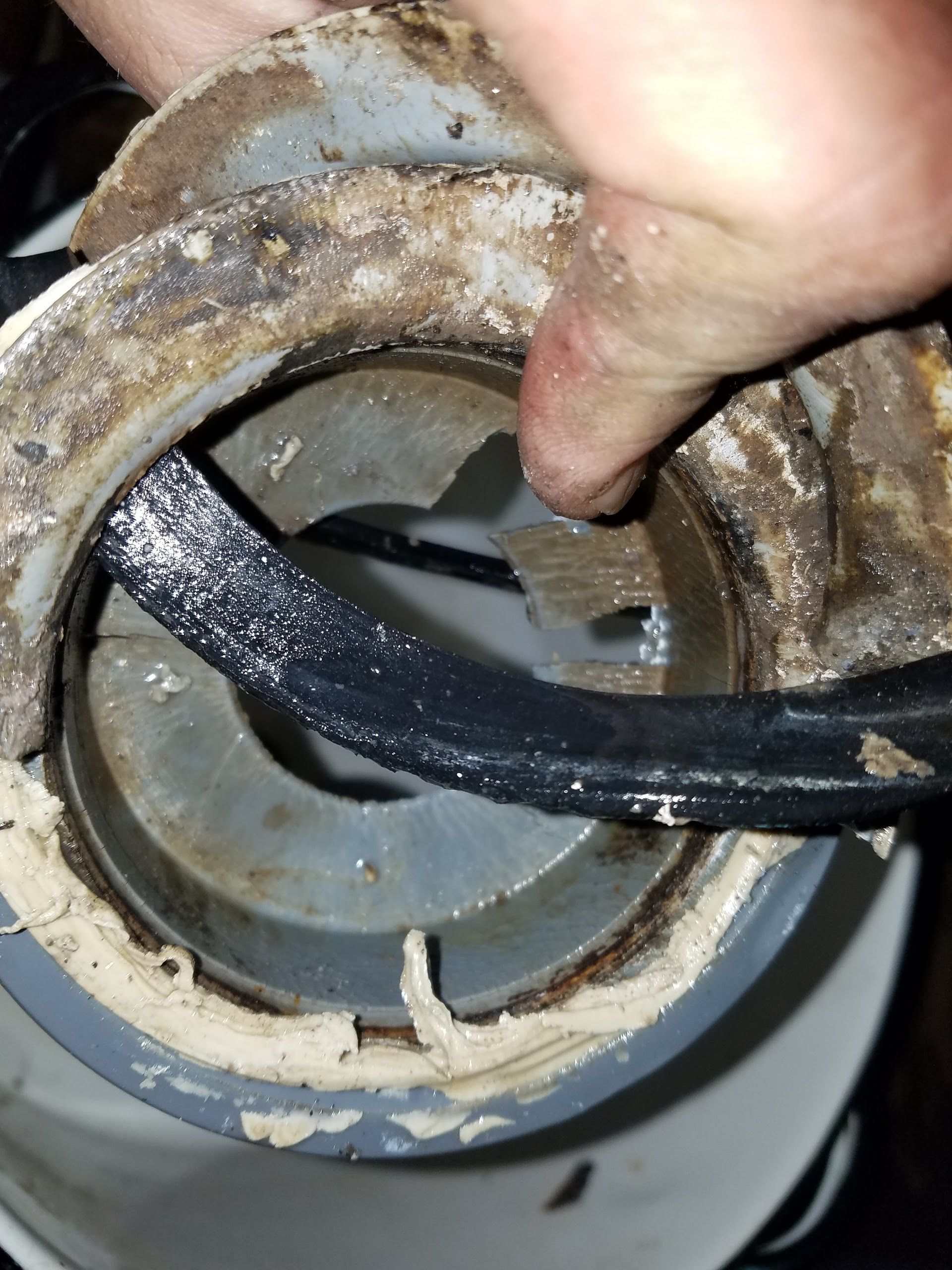 Hand points at broken part of a white plumbing fitting with black rubber seal, covered in grime.