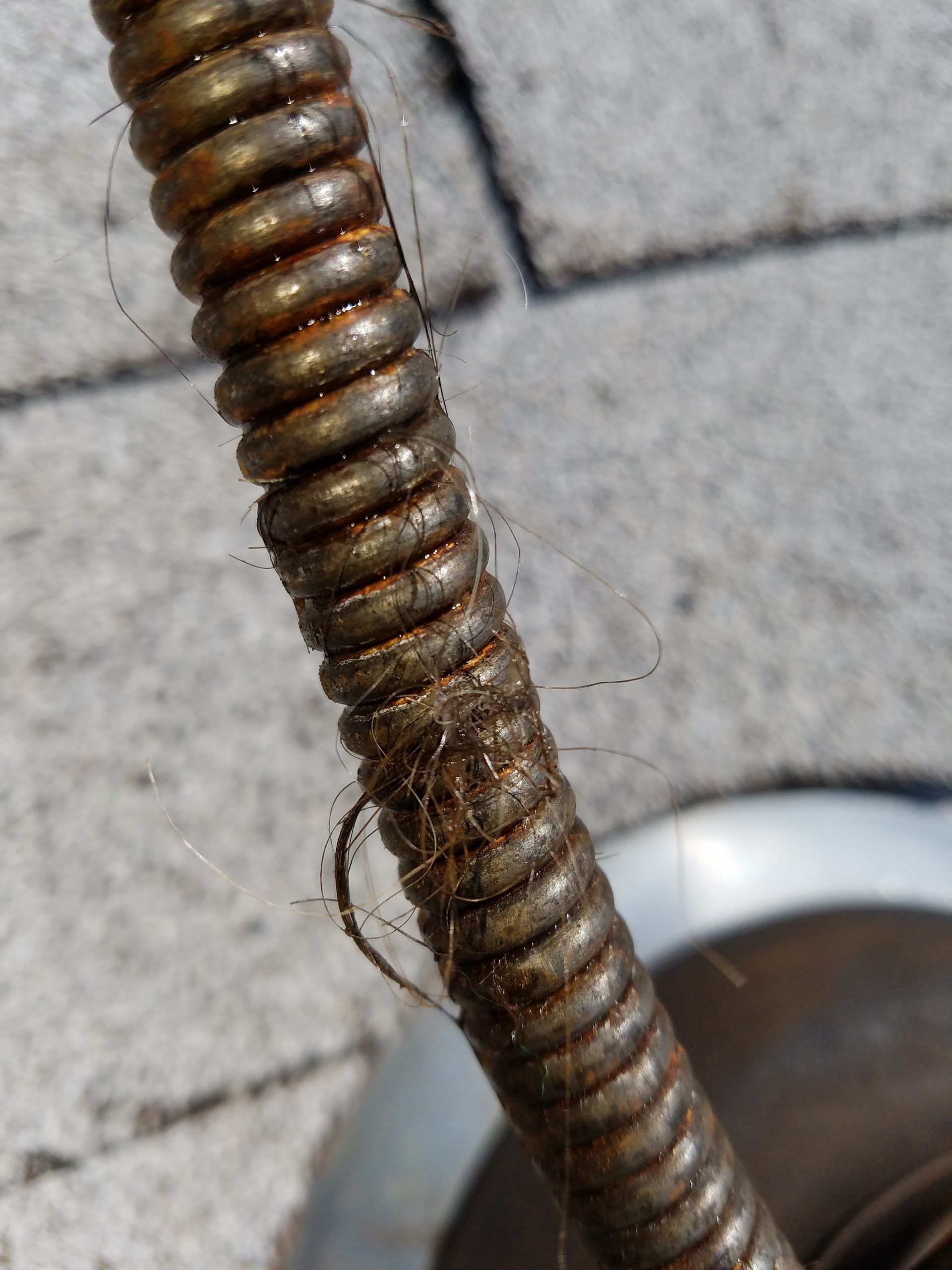 Rusty, coiled drain snake with debris, resting on a gray surface near a metal pipe.