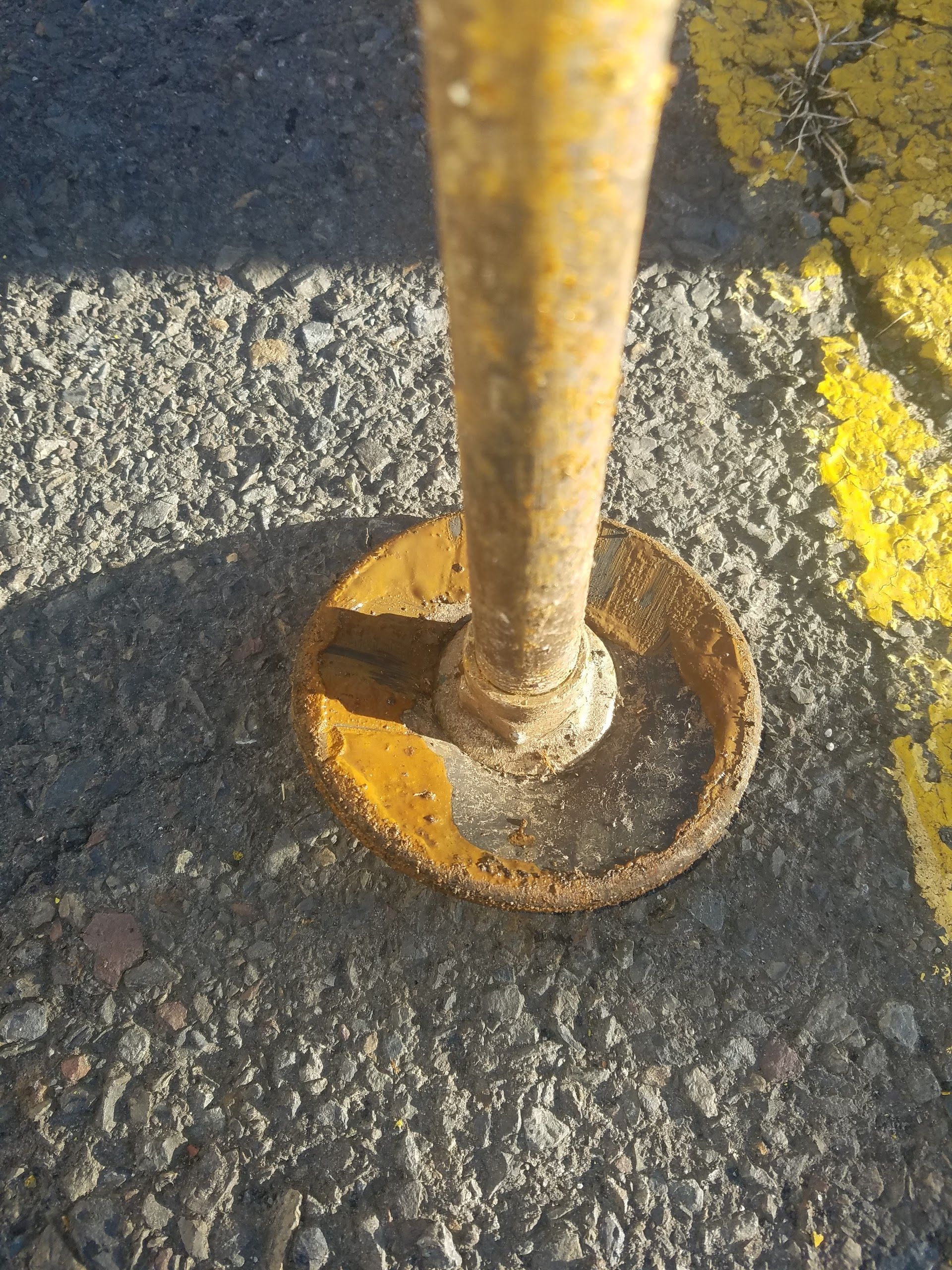 Rusty yellow metal pole base on gray asphalt, with a yellow line visible.