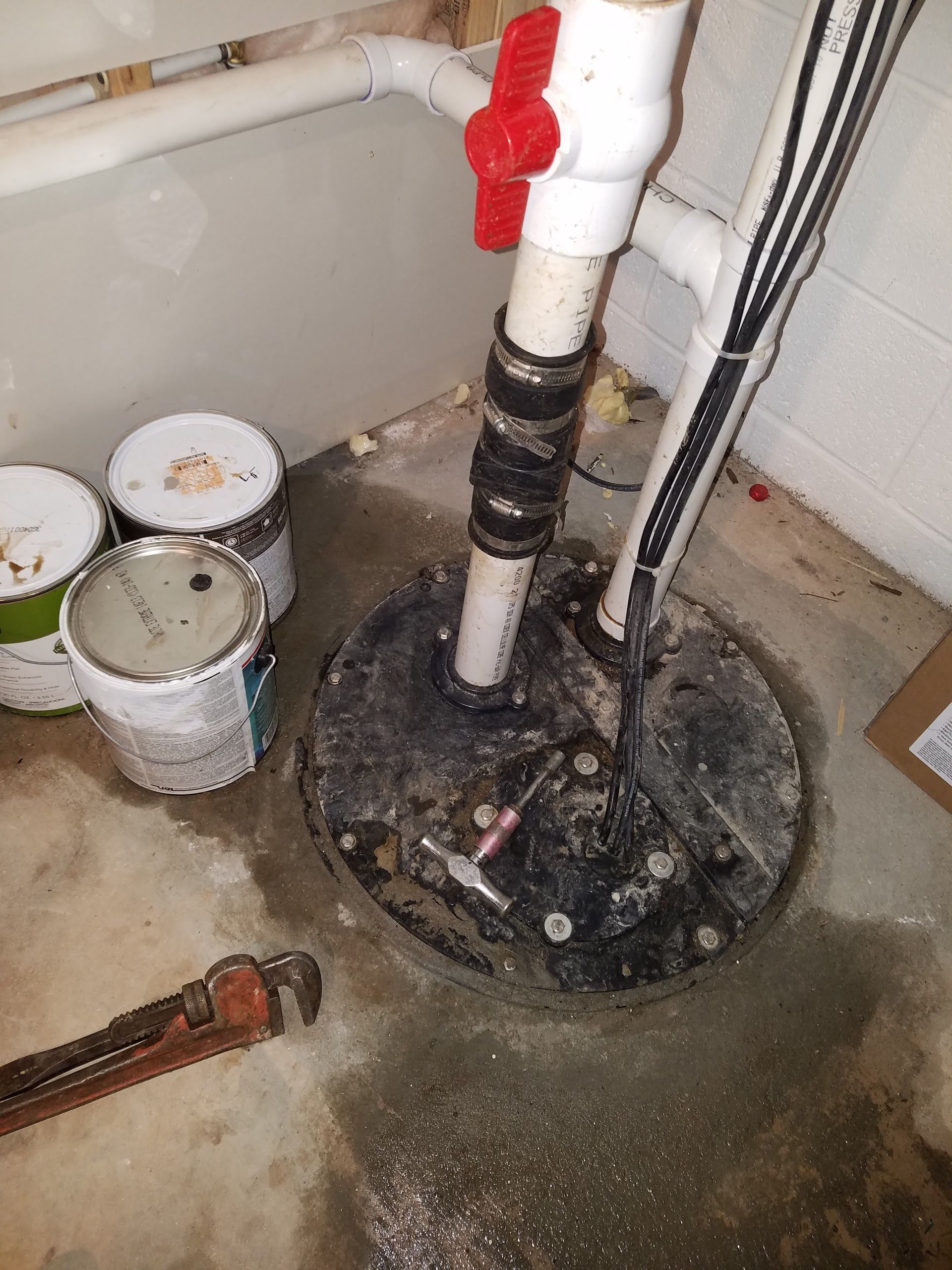 Sump pump in a basement, with white pipes, a red valve, and paint cans.