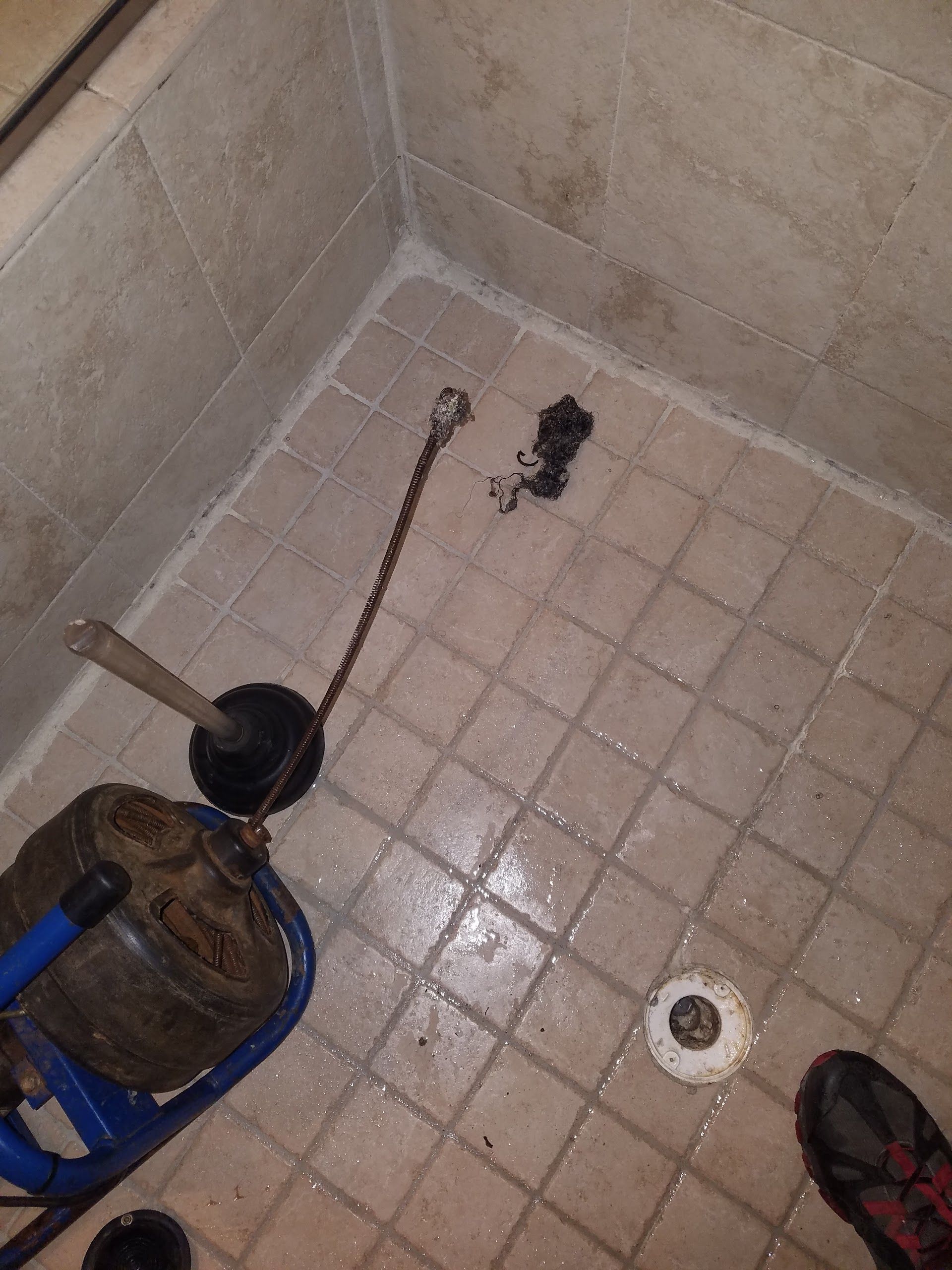 Shower stall with a plunger, snake, and debris; tiles are tan.