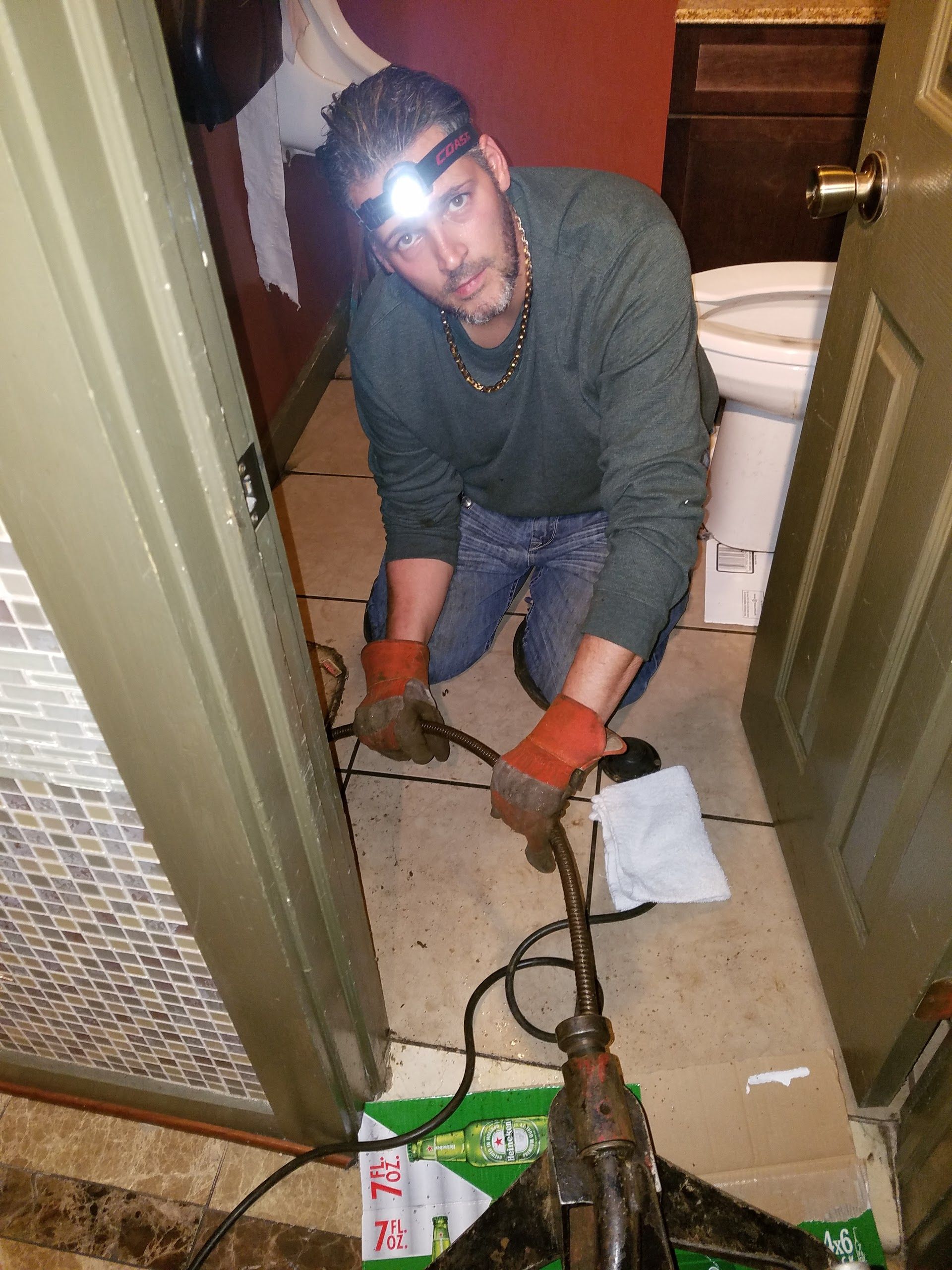 Man in bathroom, using plumbing snake, wearing headlamp, jeans, and orange gloves.