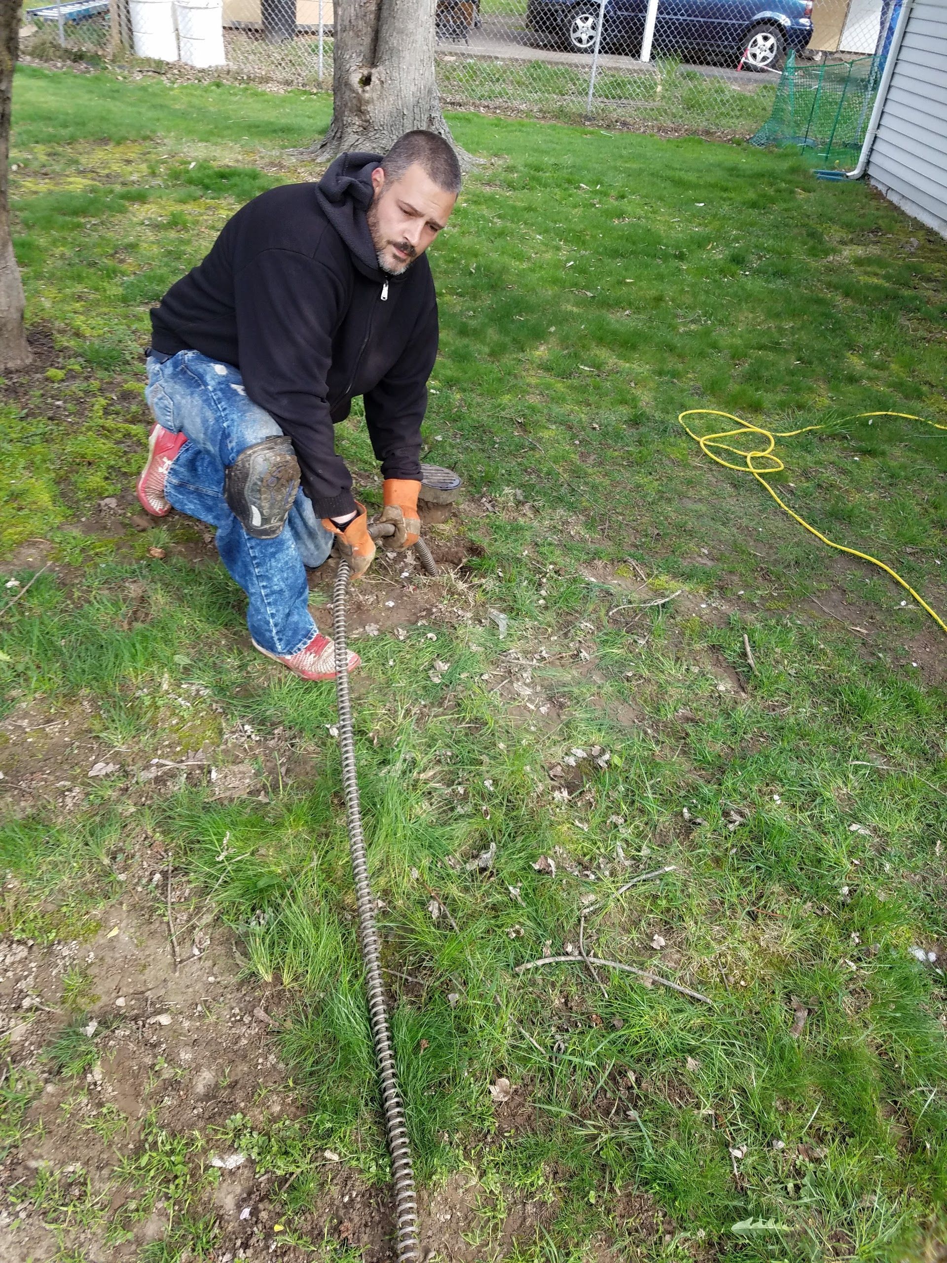 Man kneeling in grass, pulling a chain. He wears a black hoodie, blue jeans, and orange gloves.