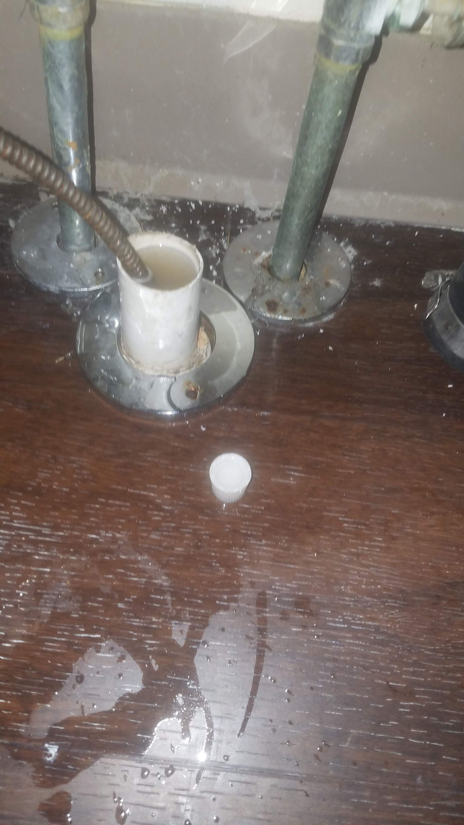 Plumbing under a sink; a drain snake is inserted into the drain. Water is on the dark wooden surface.