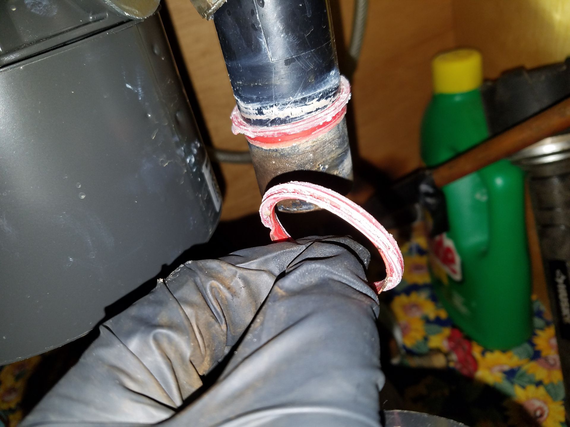 A person in black gloves holds a damaged pink gasket near a plumbing pipe connection with red sealant.