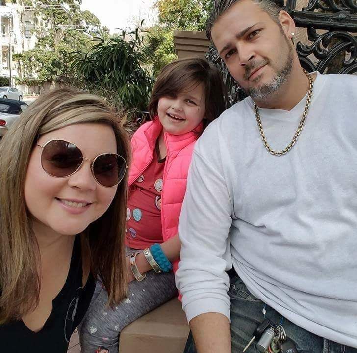 Woman, girl, and man smiling outdoors. Woman wears sunglasses, girl a pink vest, man has facial hair.