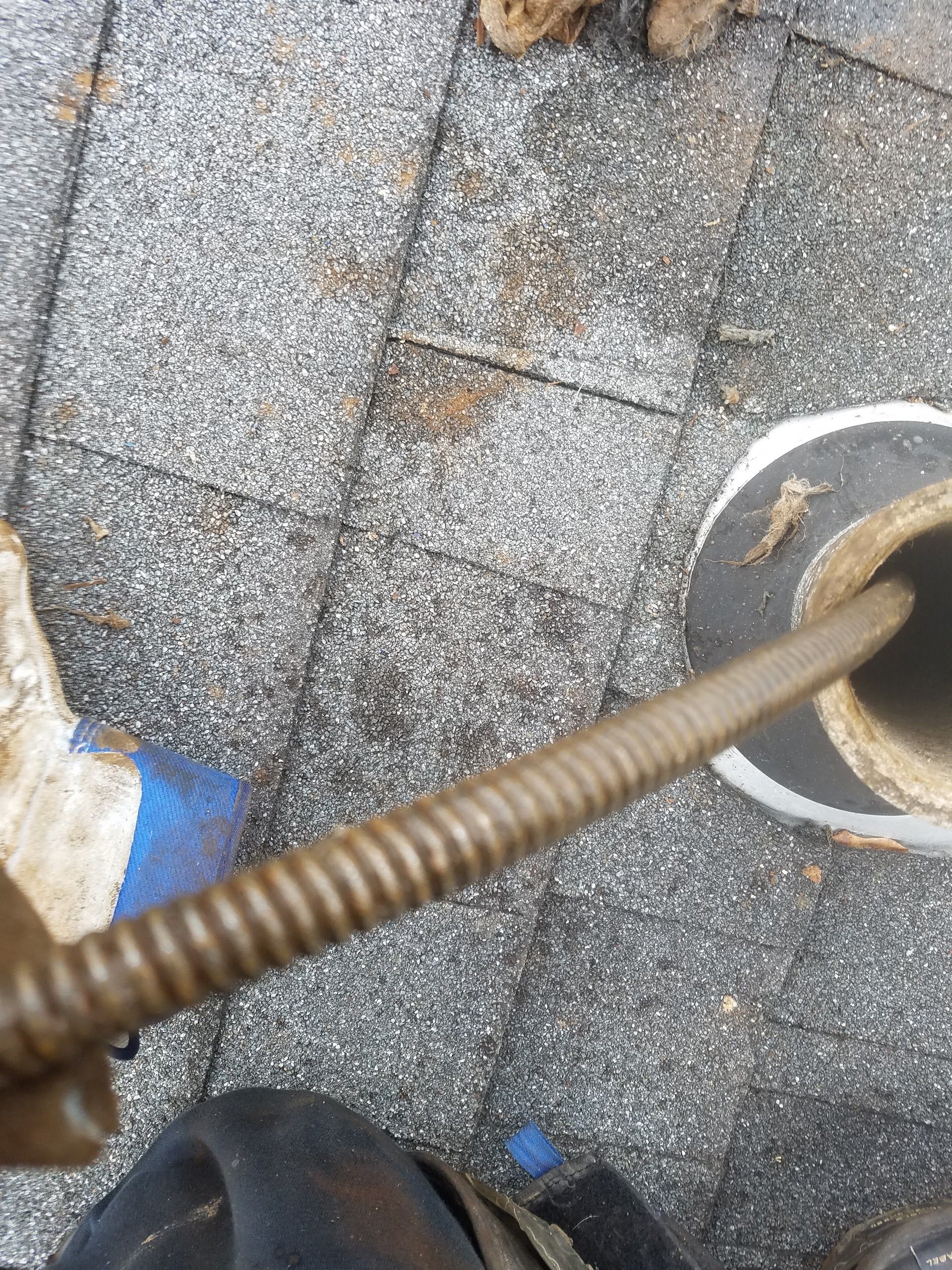 A person's hand using a snake tool to unclog a pipe on a shingled roof.