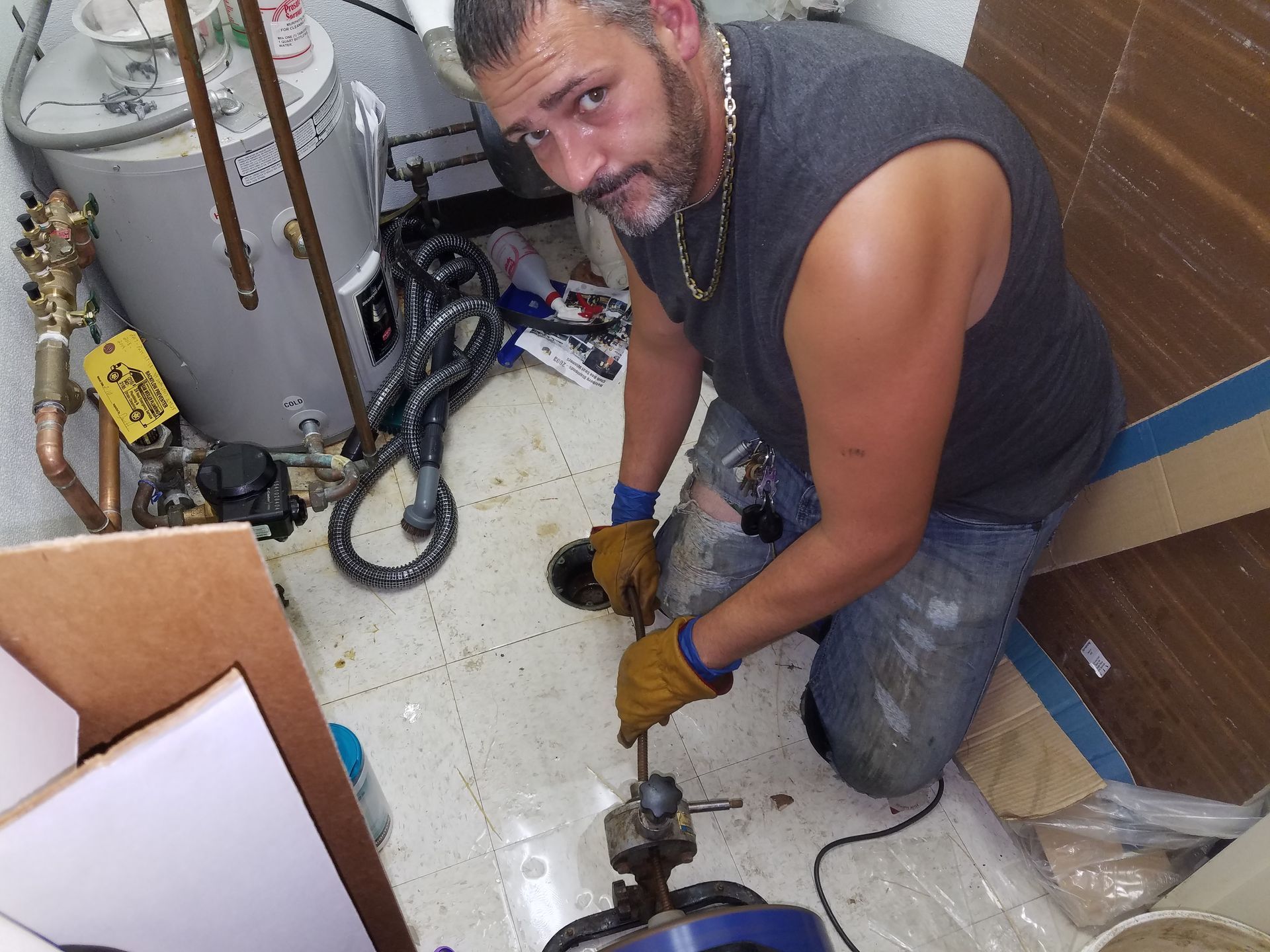 A man kneels, using a tool in a utility room. He wears work gloves, a tank top, and looks at the camera.