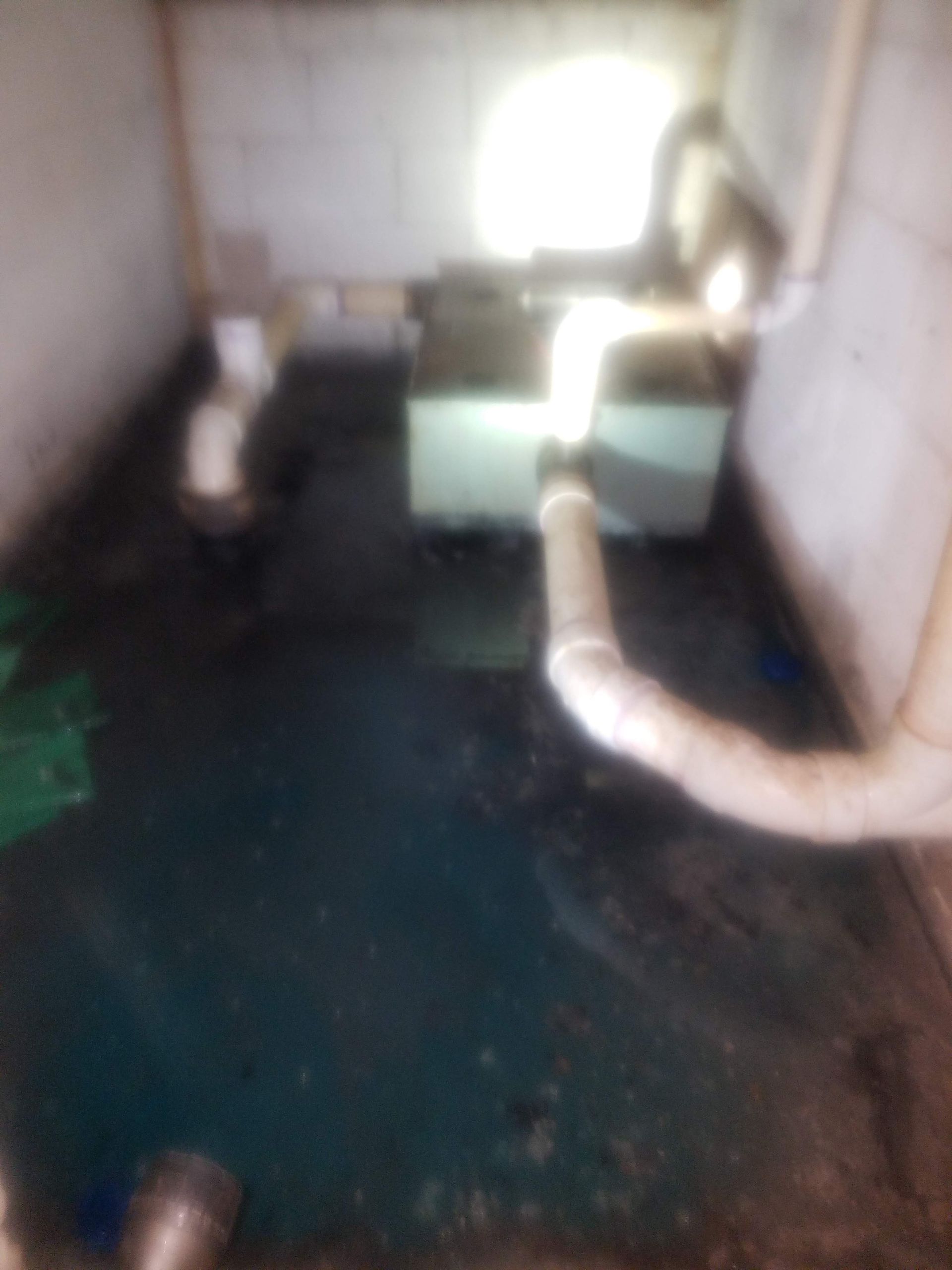 Dark room with plumbing. White pipes lead to a rectangular, light green box. Dark floor, bright light source.