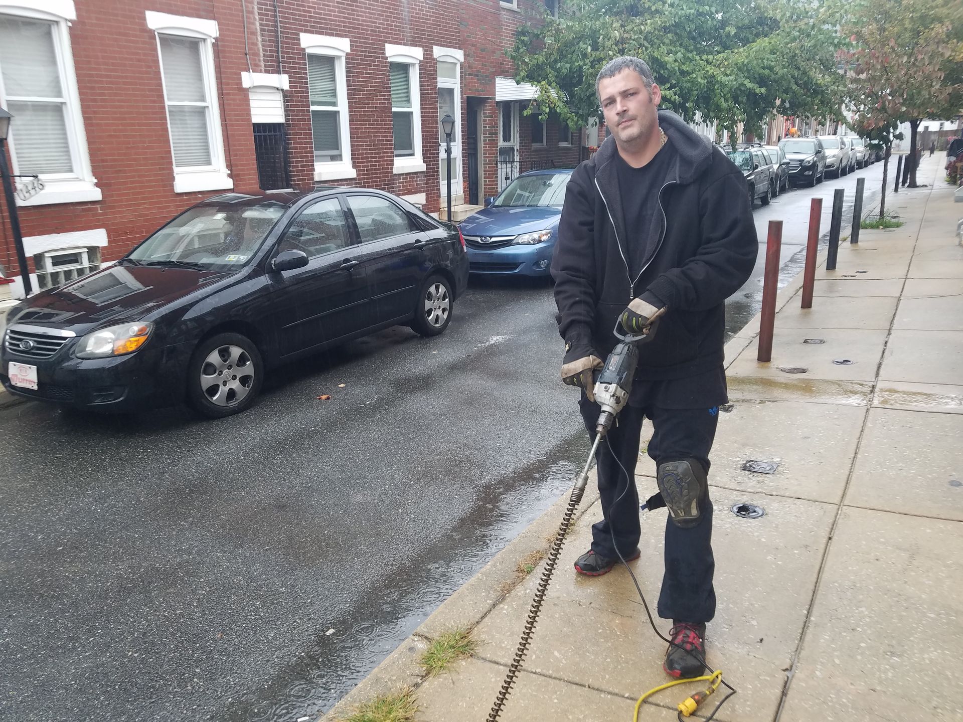 Man jackhammering sidewalk next to a street of parked cars, in front of brick buildings.