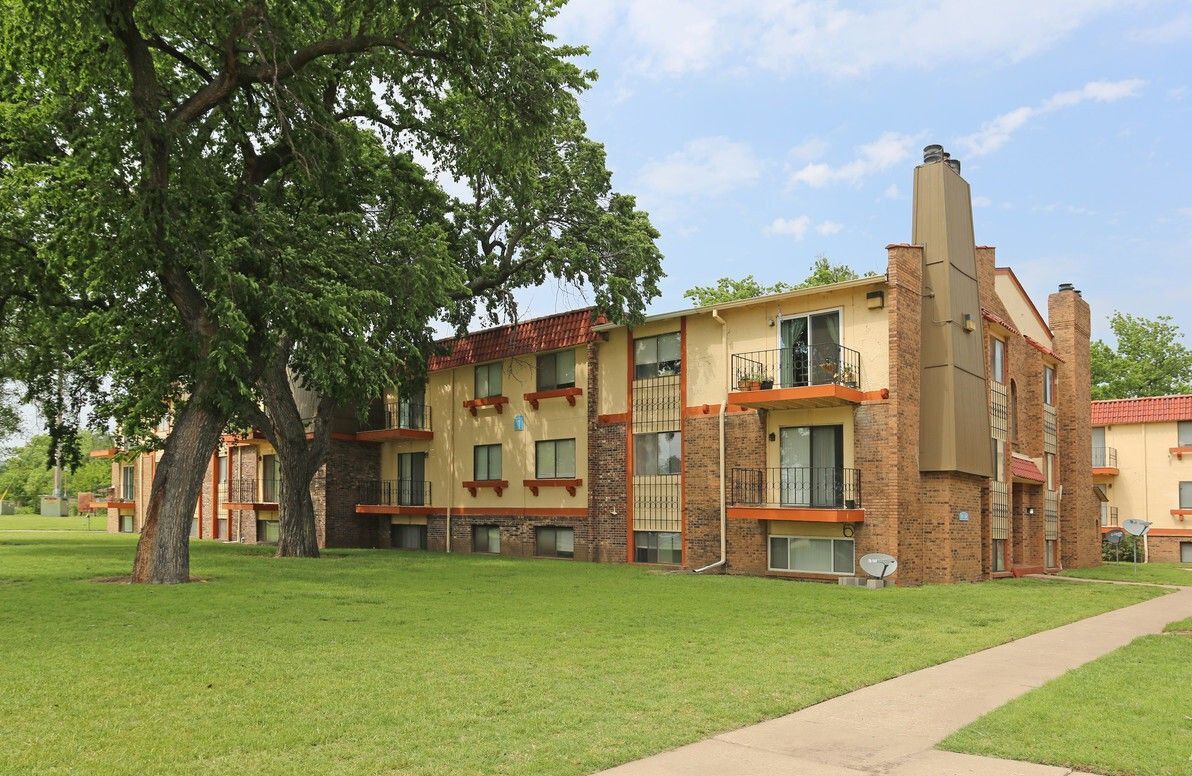 Apartment building with balconies and a brick chimney, under a large tree on a grassy lawn.