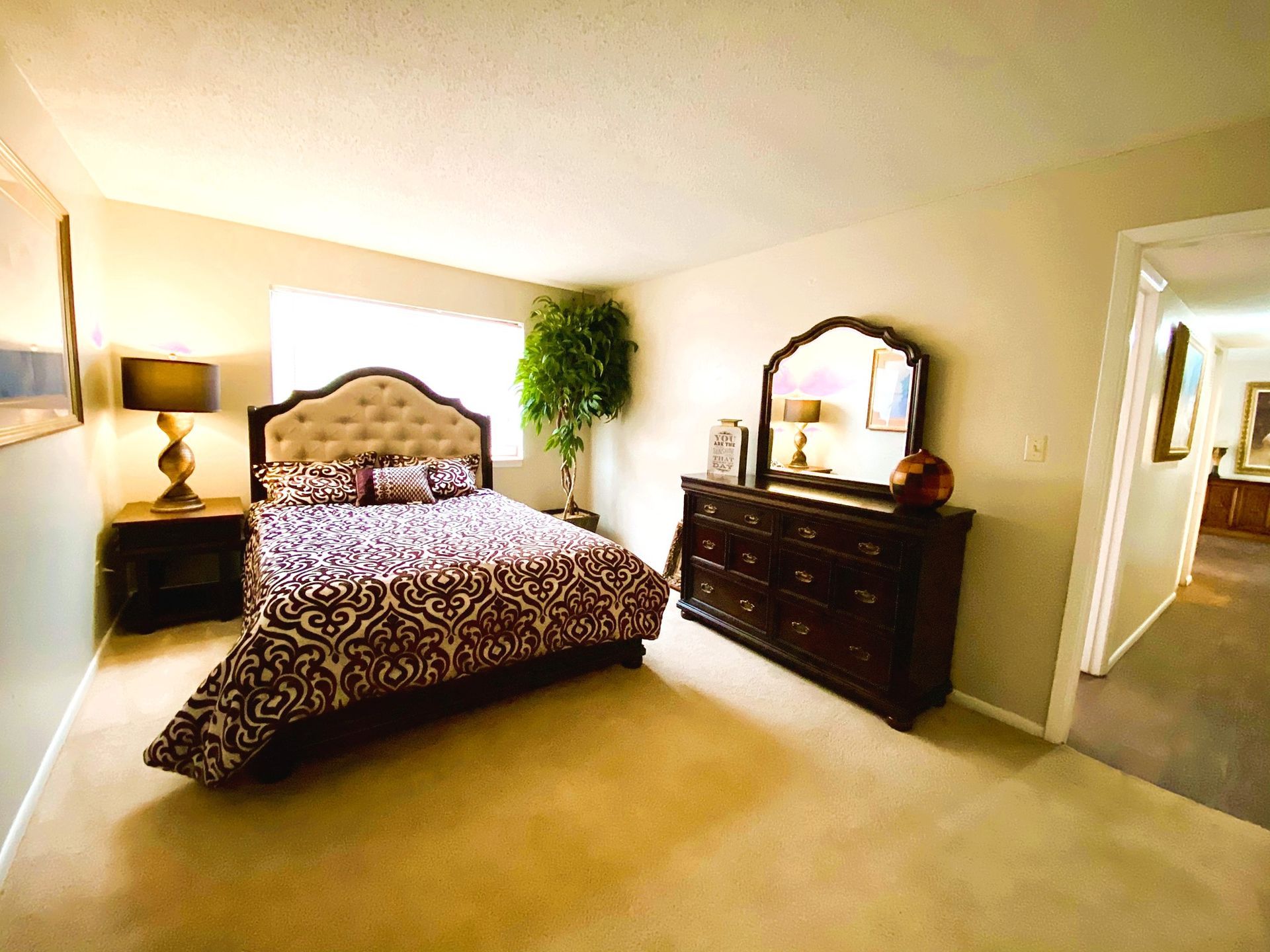 Bedroom with bed, dresser, nightstand, and tree against beige walls and carpet.