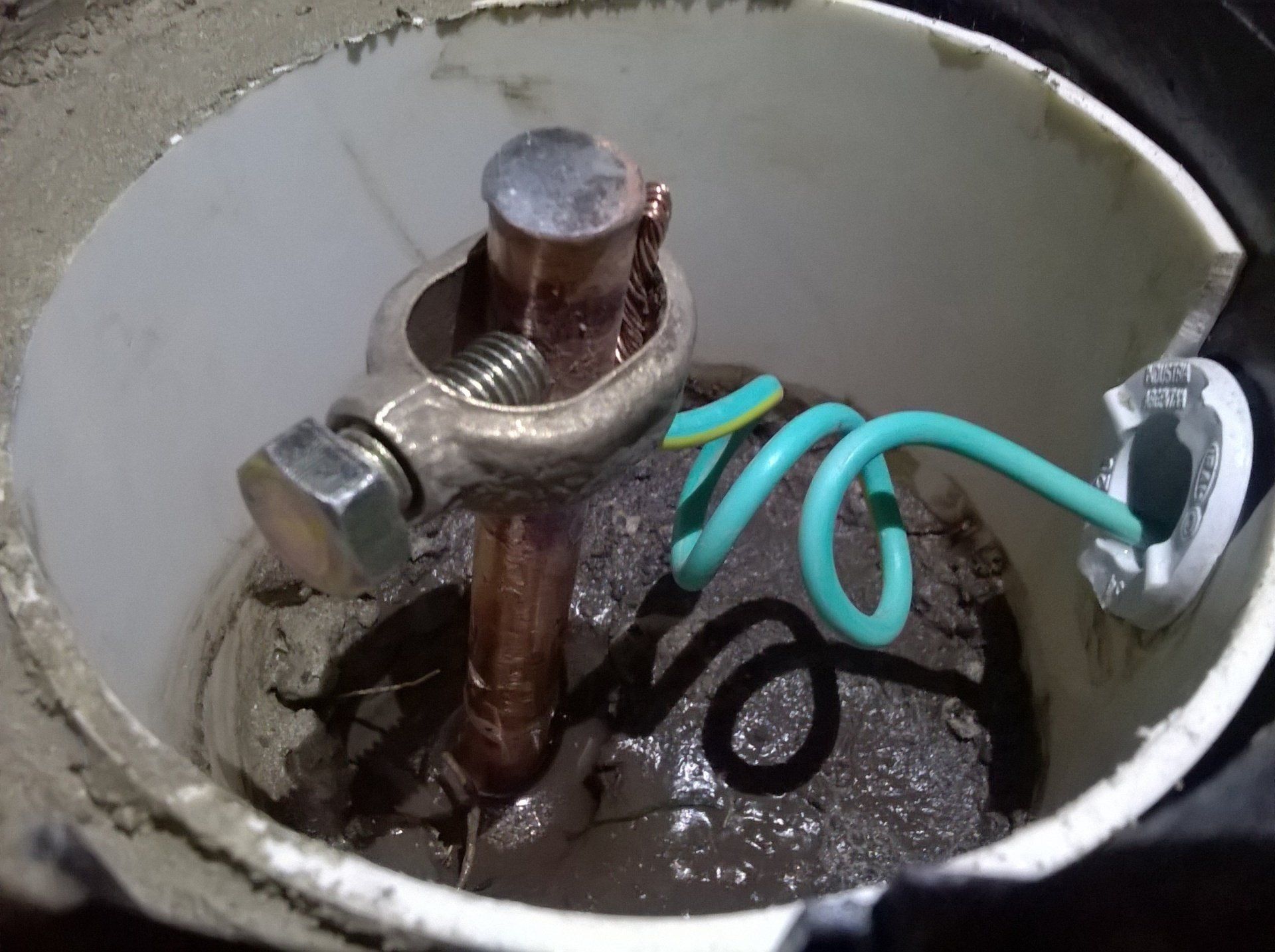 Una varilla de puesta a tierra de cobre dentro de una caja de acceso con una abrazadera metálica y un cable de puesta a tierra verde y amarillo en forma de bucle.