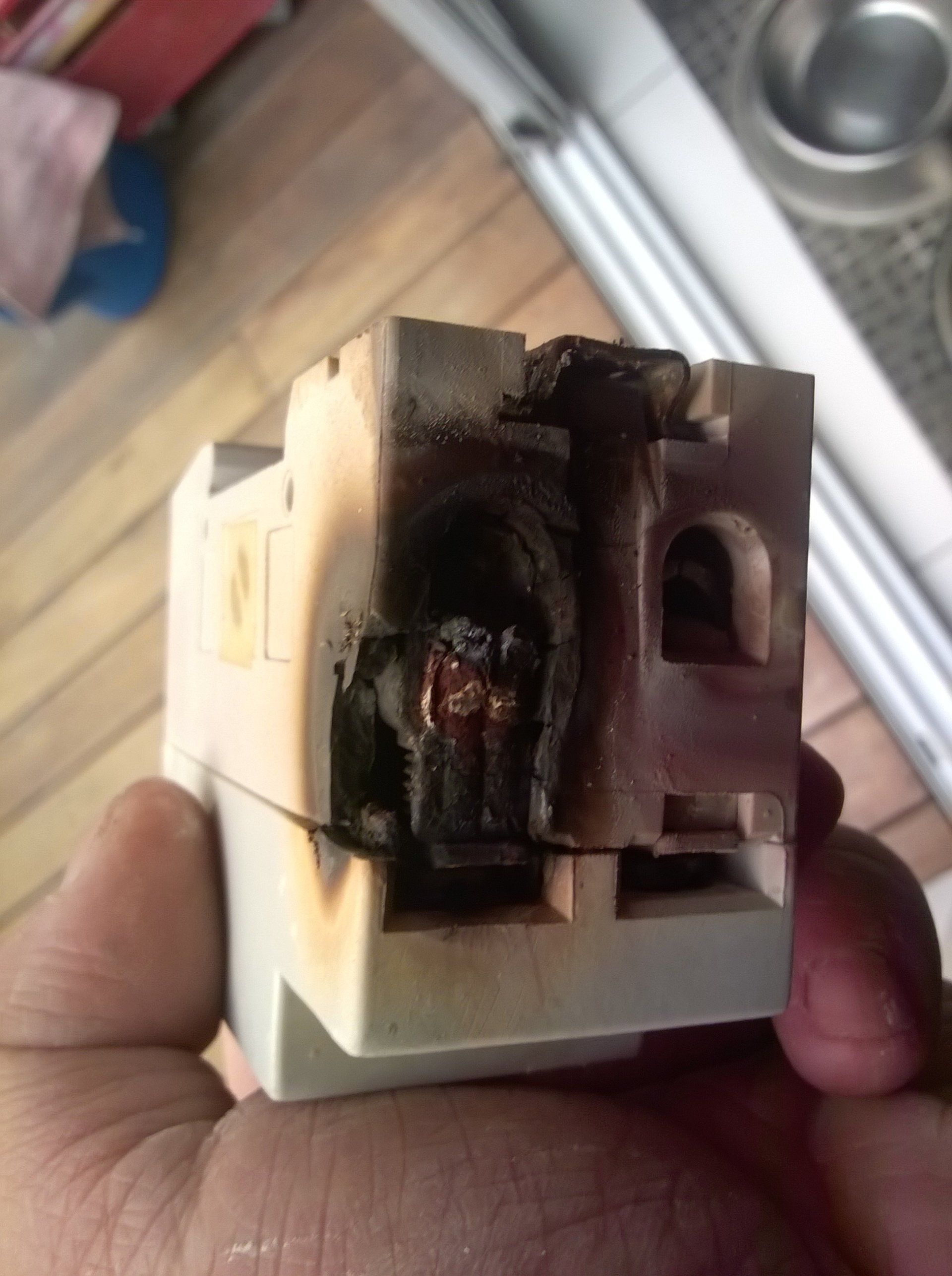 Una mano sostiene un interruptor automático eléctrico gravemente quemado y derretido, mostrando el plástico chamuscado y los componentes internos dañados.