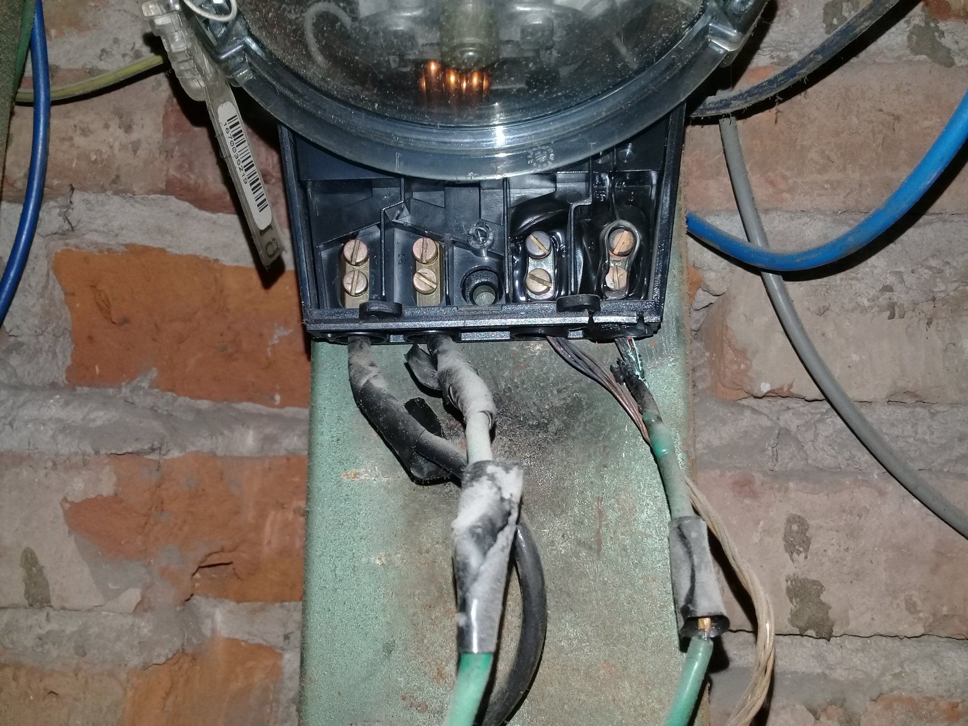 Un contador eléctrico montado en una pared de ladrillo, con cables visibles y conexiones encintadas en los terminales.
