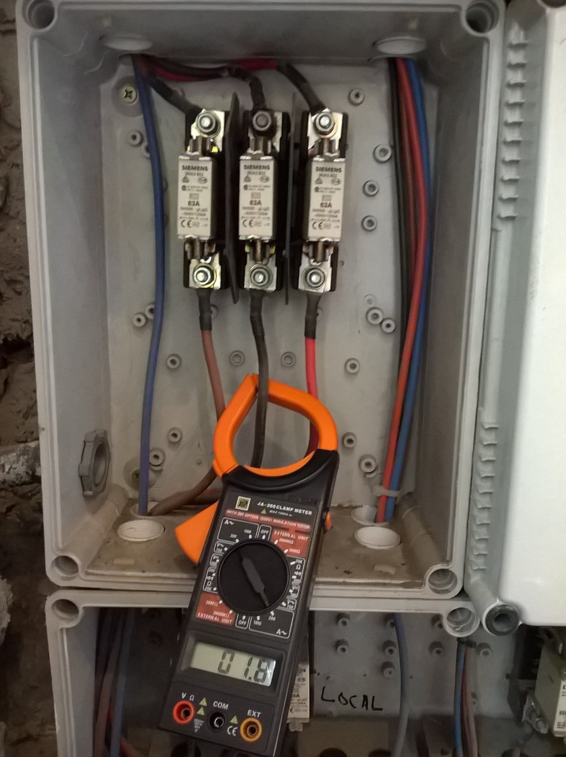 Un amperímetro de pinza mide la corriente eléctrica en un cable conectado a un bloque de fusibles dentro de una caja de conexiones gris abierta.