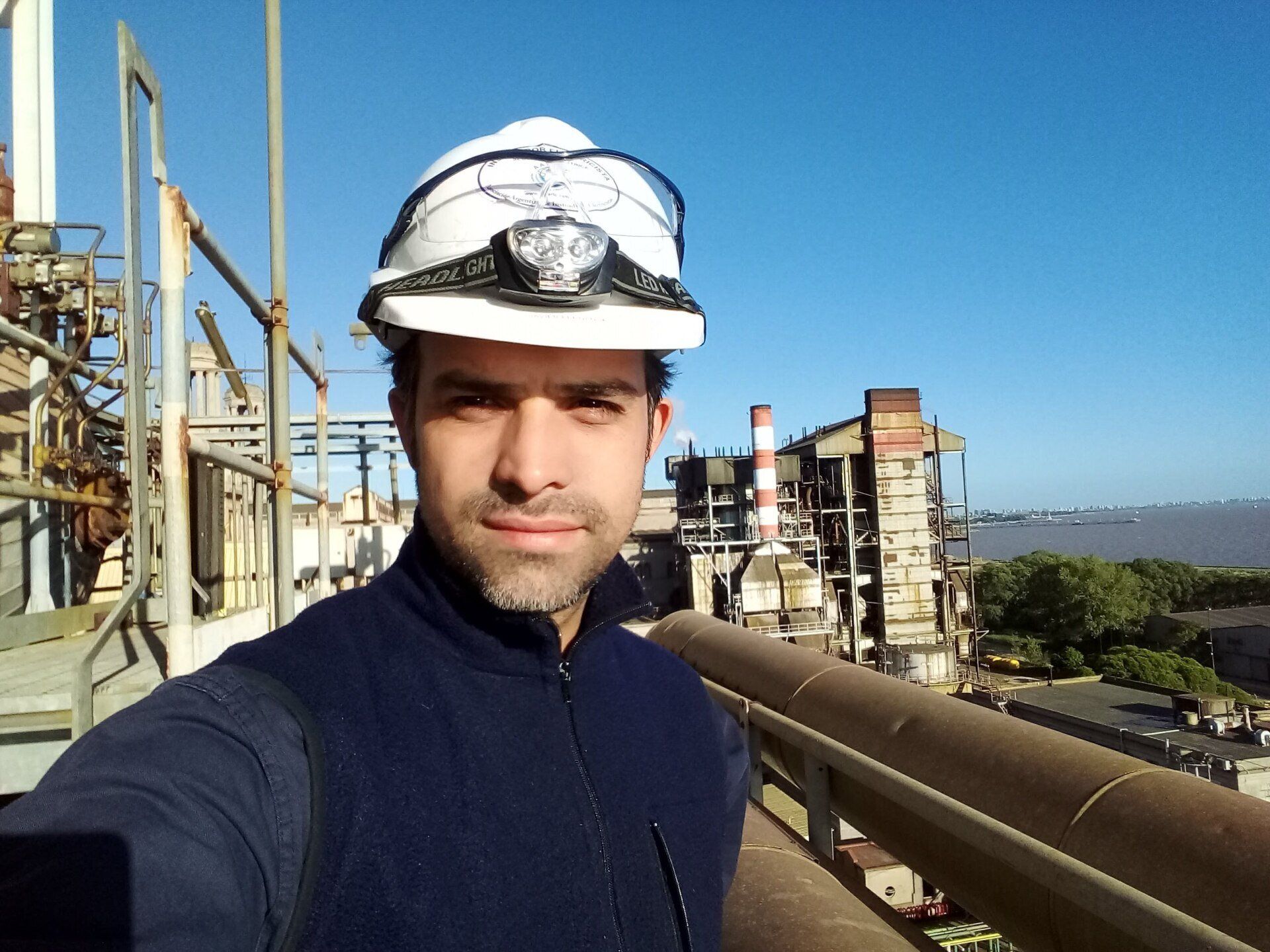 Un trabajador con casco blanco y chaleco oscuro está de pie en un balcón con vistas a una instalación industrial y al agua.