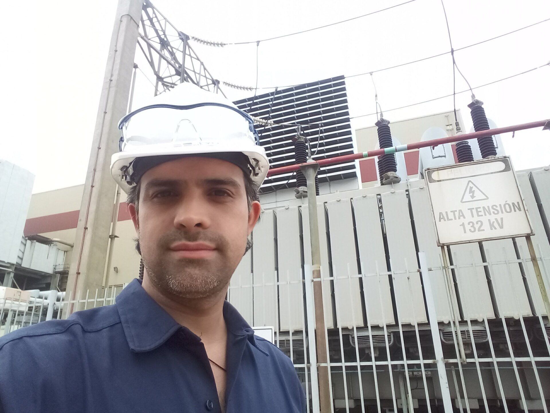 Una persona con casco y gafas protectoras se encuentra de pie frente a una subestación eléctrica de alta tensión.