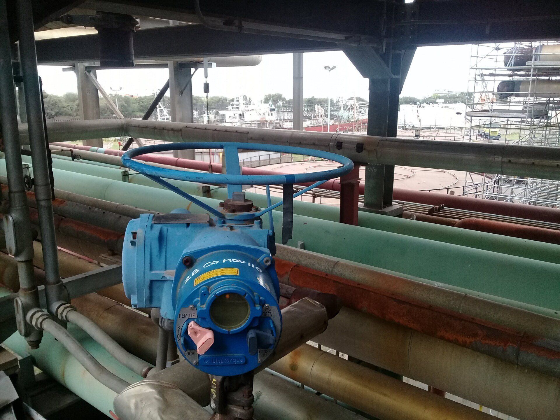 Una válvula industrial azul con volante montada sobre tuberías en una instalación, con un astillero visible al fondo.