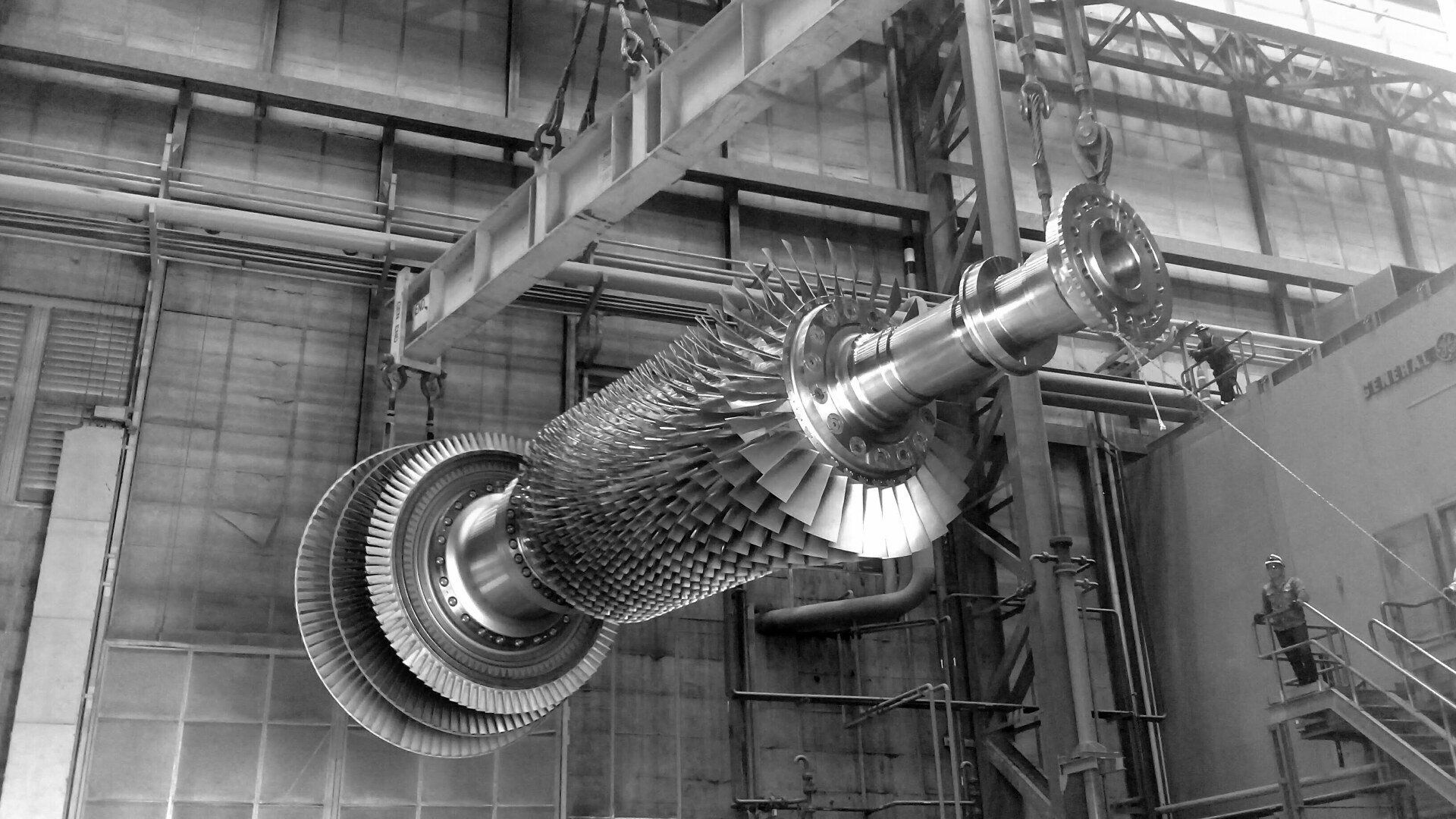 En un entorno fabril, el rotor de una gran turbina de vapor industrial cuelga de una grúa aérea.