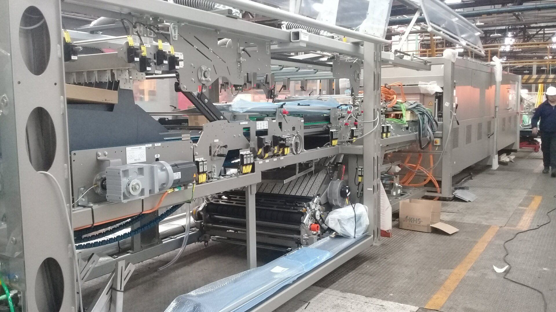 Una larga máquina de embalaje industrial con estructura metálica y cintas transportadoras en un almacén, con un trabajador cerca.
