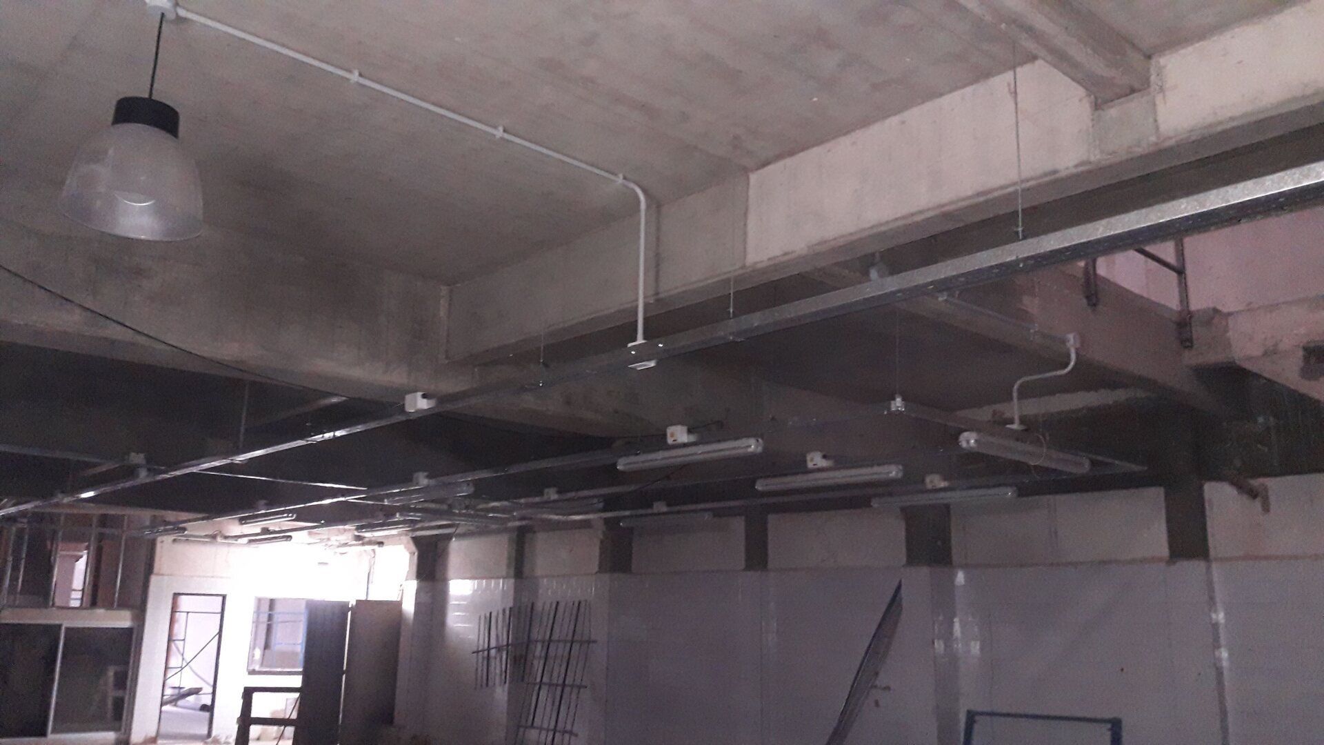 Una obra en construcción con techos de hormigón visto, bandejas metálicas portacables, tuberías y luminarias colgantes.