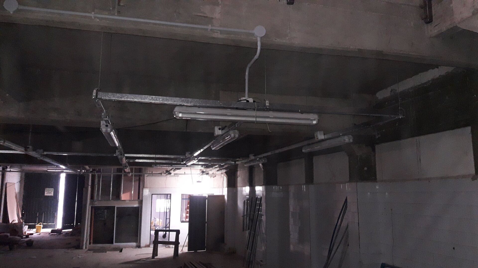 Una obra en construcción con techo sin terminar, hormigón visto, luces fluorescentes colgantes y conductos metálicos.