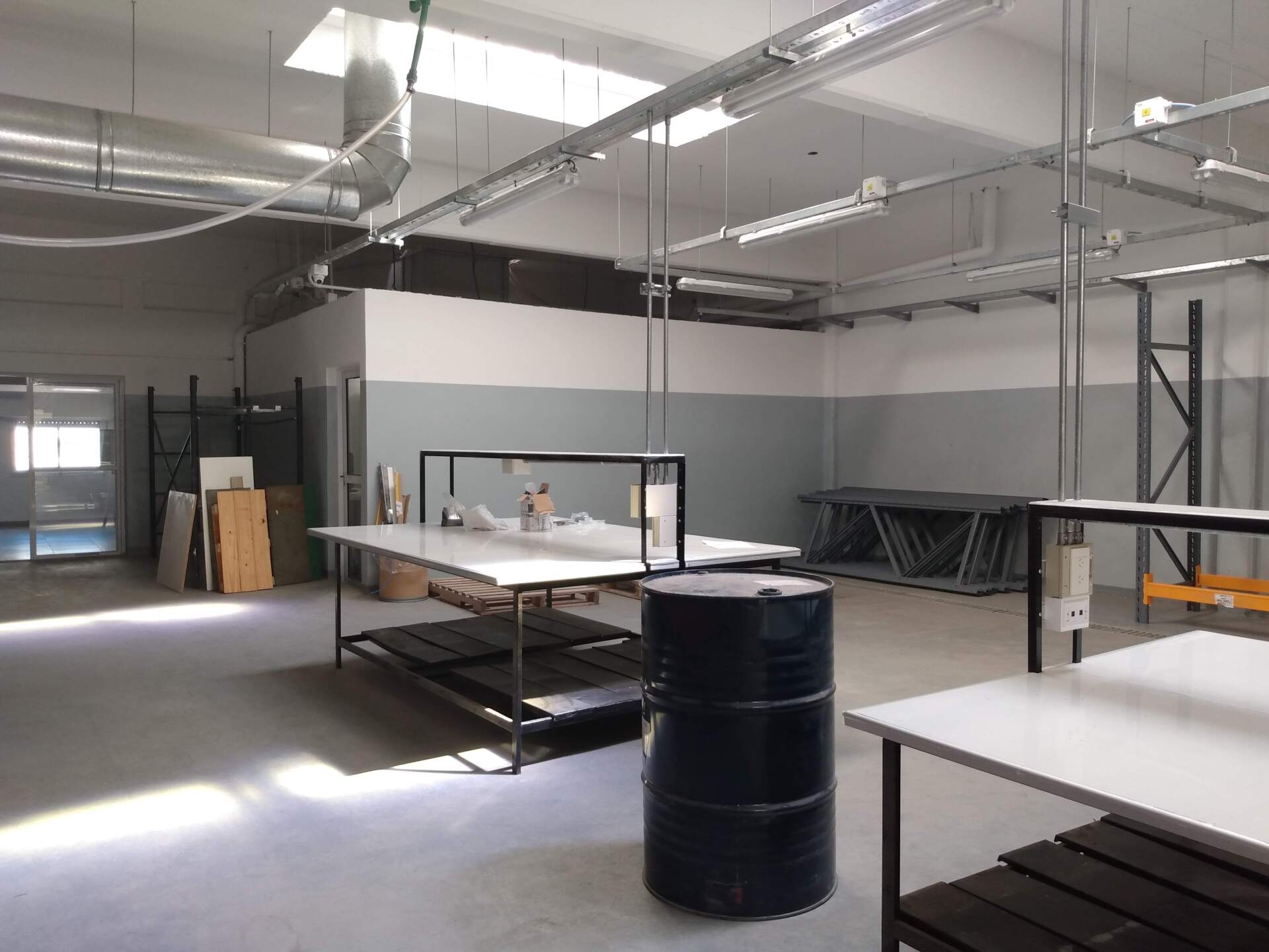 Un taller industrial espacioso con paredes blancas y grises, mesas de trabajo metálicas, un barril negro e iluminación cenital.
