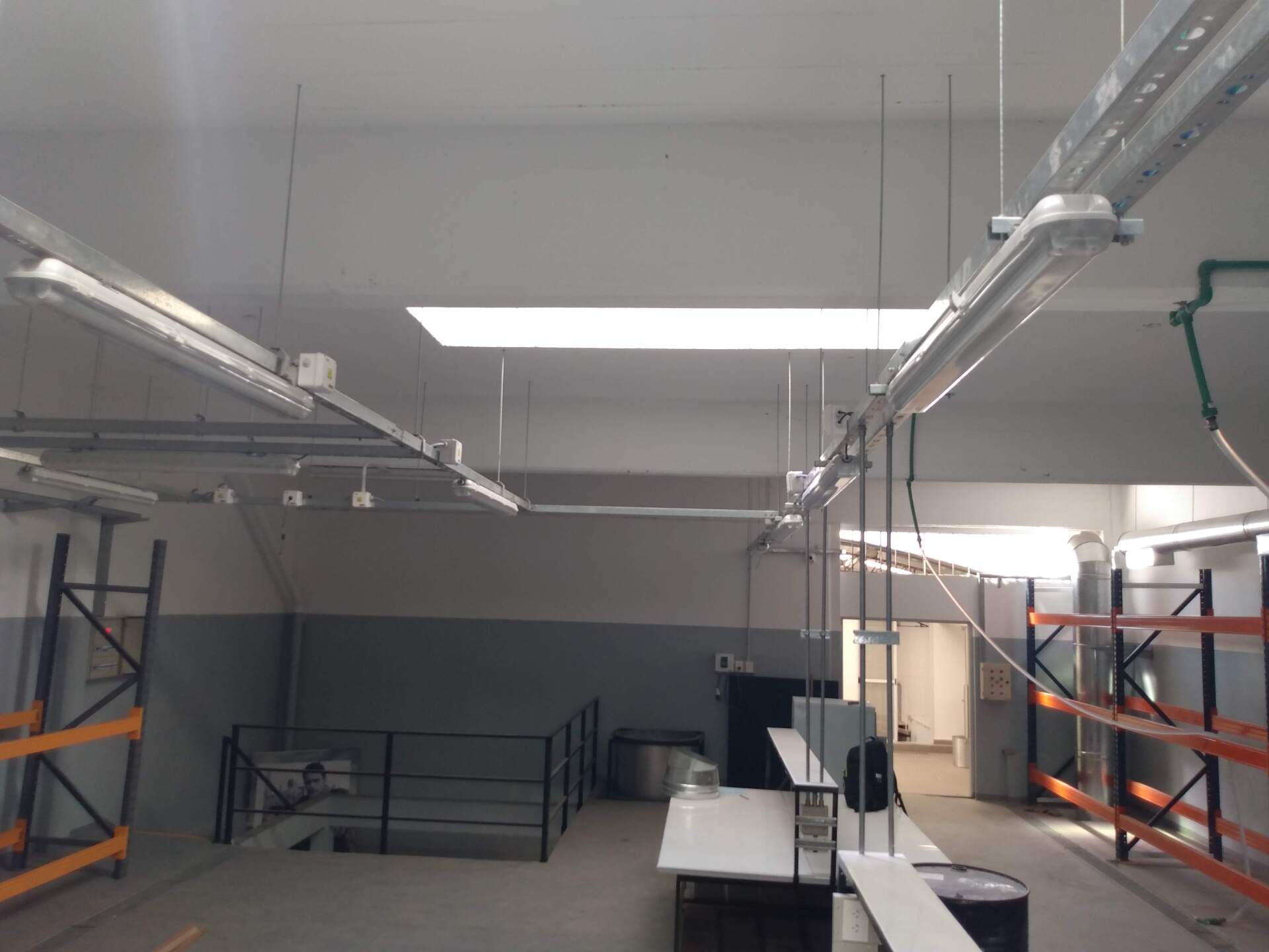 Un espacio industrial interior con luces fluorescentes colgantes, estanterías y una plataforma central con barandilla metálica.